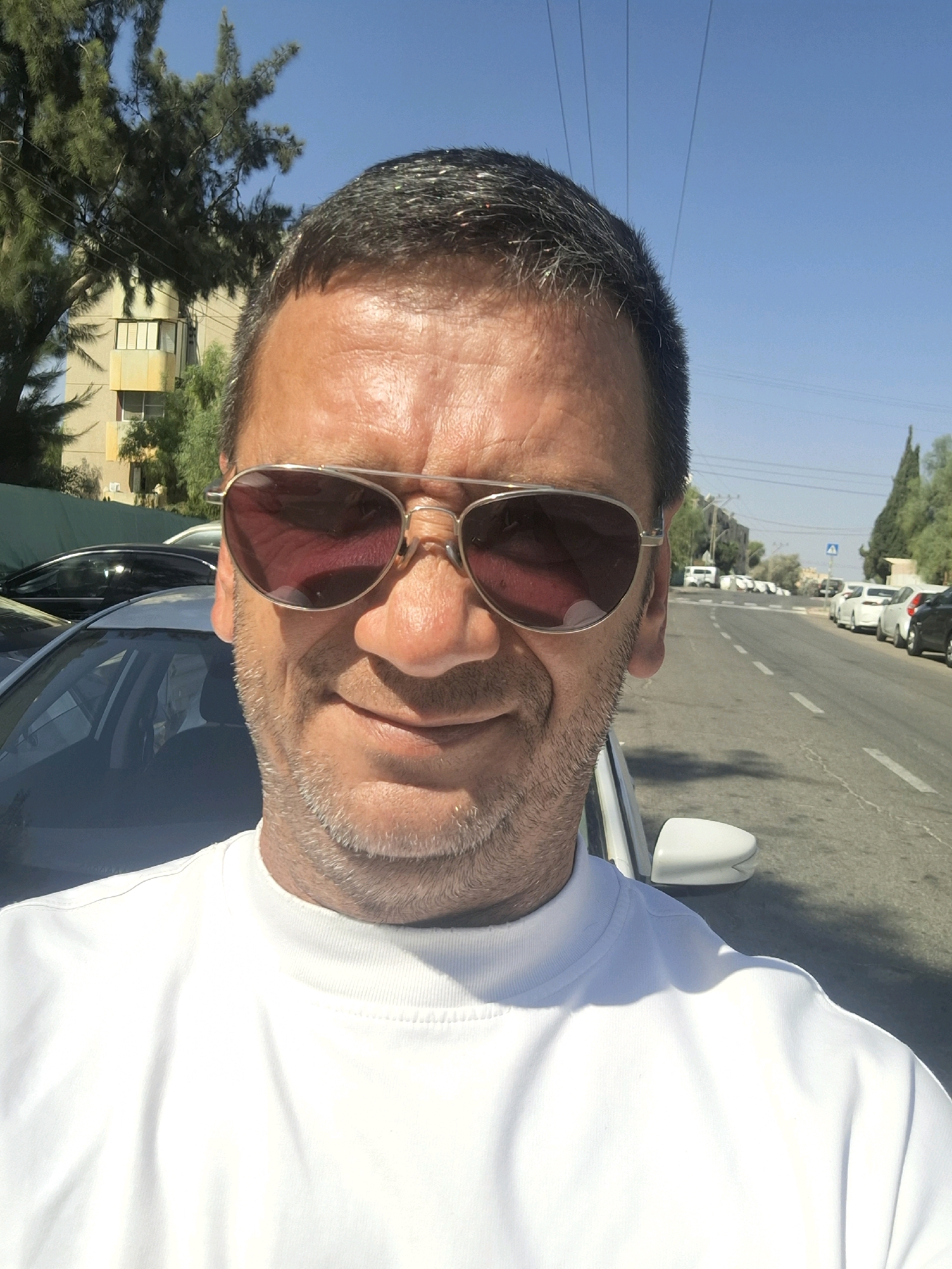 A man wearing sunglasses and a white t-shirt takes a selfie on a street in Be'er-Sheva, Israel. The man has short, dark hair, a light beard, and is looking directly at the camera. Behind him, a street lined with parked cars and trees is visible. A yellow building and telephone poles are also present in the background. The sky is clear and blue.