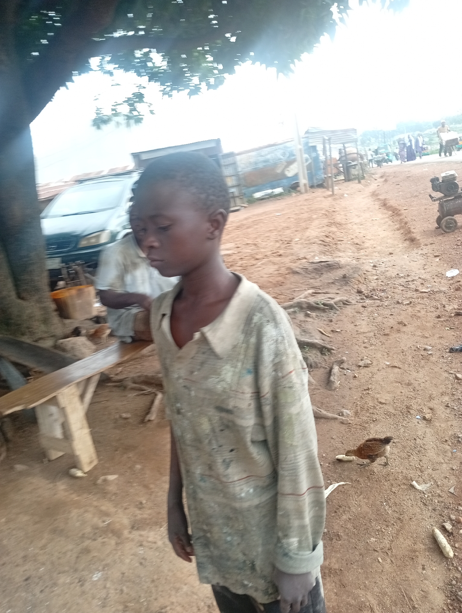 A medium shot shows a young boy standing in a dusty outdoor environment, possibly a market or roadside stall in Rafin Par, Nigeria. He is wearing a faded, light-colored long-sleeved shirt with a patterned design. His face is turned to his right, with his gaze directed downwards. Behind him, to his left, a portion of a dark tree trunk is visible. Further back, a light-colored vehicle with its front visible is parked. To the right of the boy, a small brown chicken is pecking at a piece of corn on the ground. The ground is composed of reddish-brown dirt and scattered debris. In the background, there are various structures, including what appears to be a stall with a corrugated metal roof, and other indistinct figures and objects, suggesting a populated area. The lighting indicates it is daytime, with a bright, hazy sky.