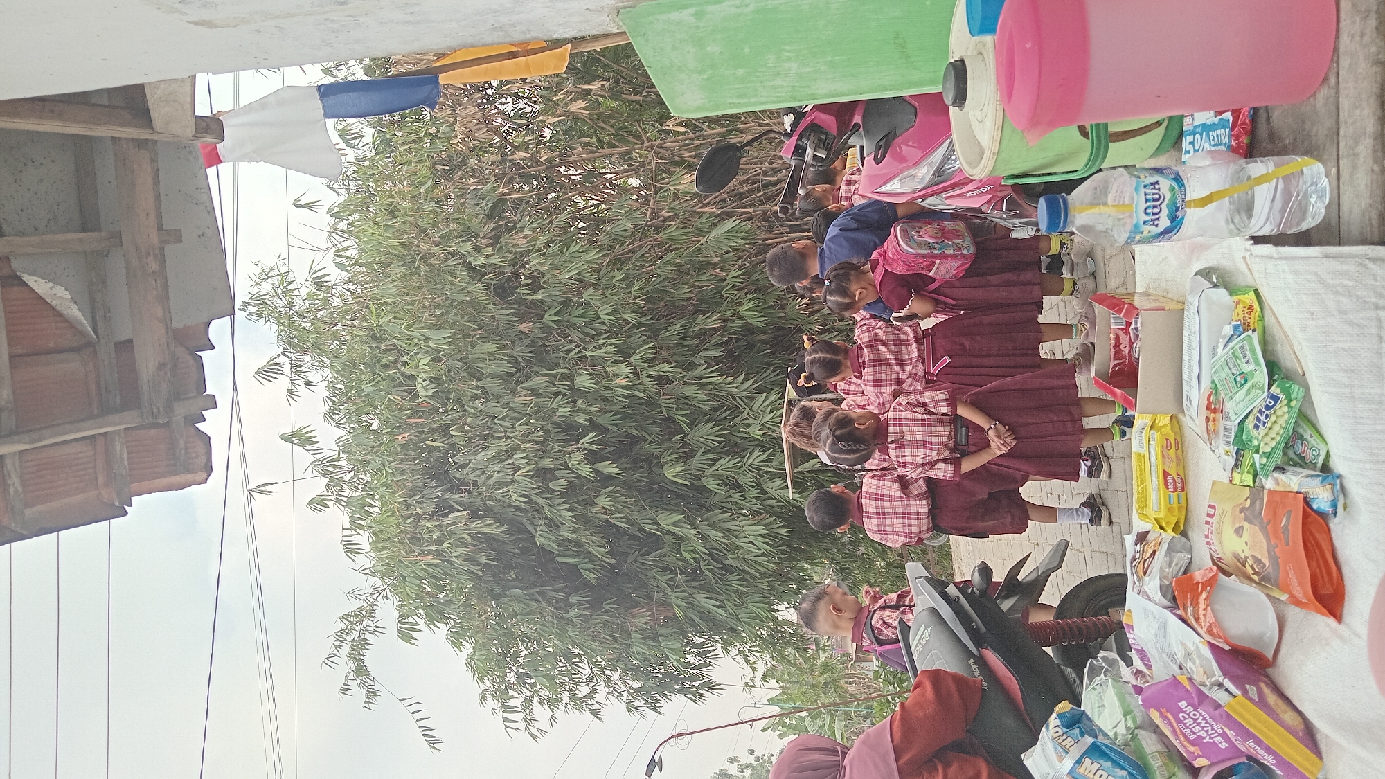 In Kedungwaru, Indonesia, a group of young students, dressed in red and white plaid uniforms with maroon skirts and shorts, are gathered outside. They are standing in front of a dense bamboo grove. A few adults are also present, including a woman wearing a maroon hijab and a dark blue shirt. In the foreground, various snacks and drinks are spread out on a white cloth, including a bottle of Aqua water, packets of snacks, and a pink pitcher. A pink scooter is parked to the right, and a black motorcycle is to the left. The environment suggests an outdoor gathering, possibly a school event or field trip, under a cloudy sky. The structure in the background shows a rustic, weathered roof.