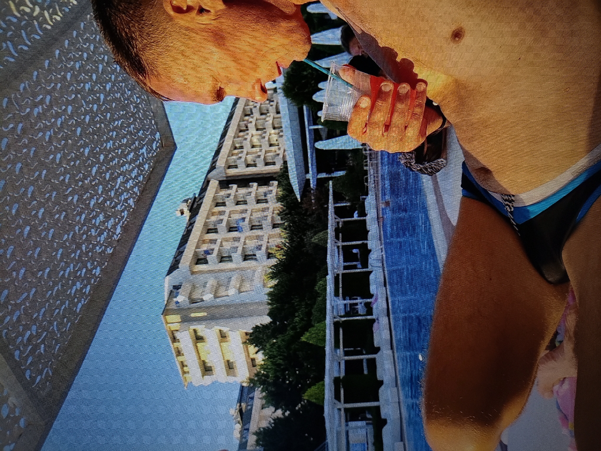 A man is relaxing under a patterned canopy near a swimming pool, holding a drink with a straw. He is wearing swim trunks. In the background, there is a large white building with balconies, trees, and a glimpse of more buildings beyond. The sky is visible above the canopy. The scene appears to be a resort or hotel. The provided crops indicate that the image is a close-up shot focusing on the man and his drink, with the background elements slightly out of focus. The question asks for the location in Tiraspol, Moldova, which cannot be determined from the image itself.