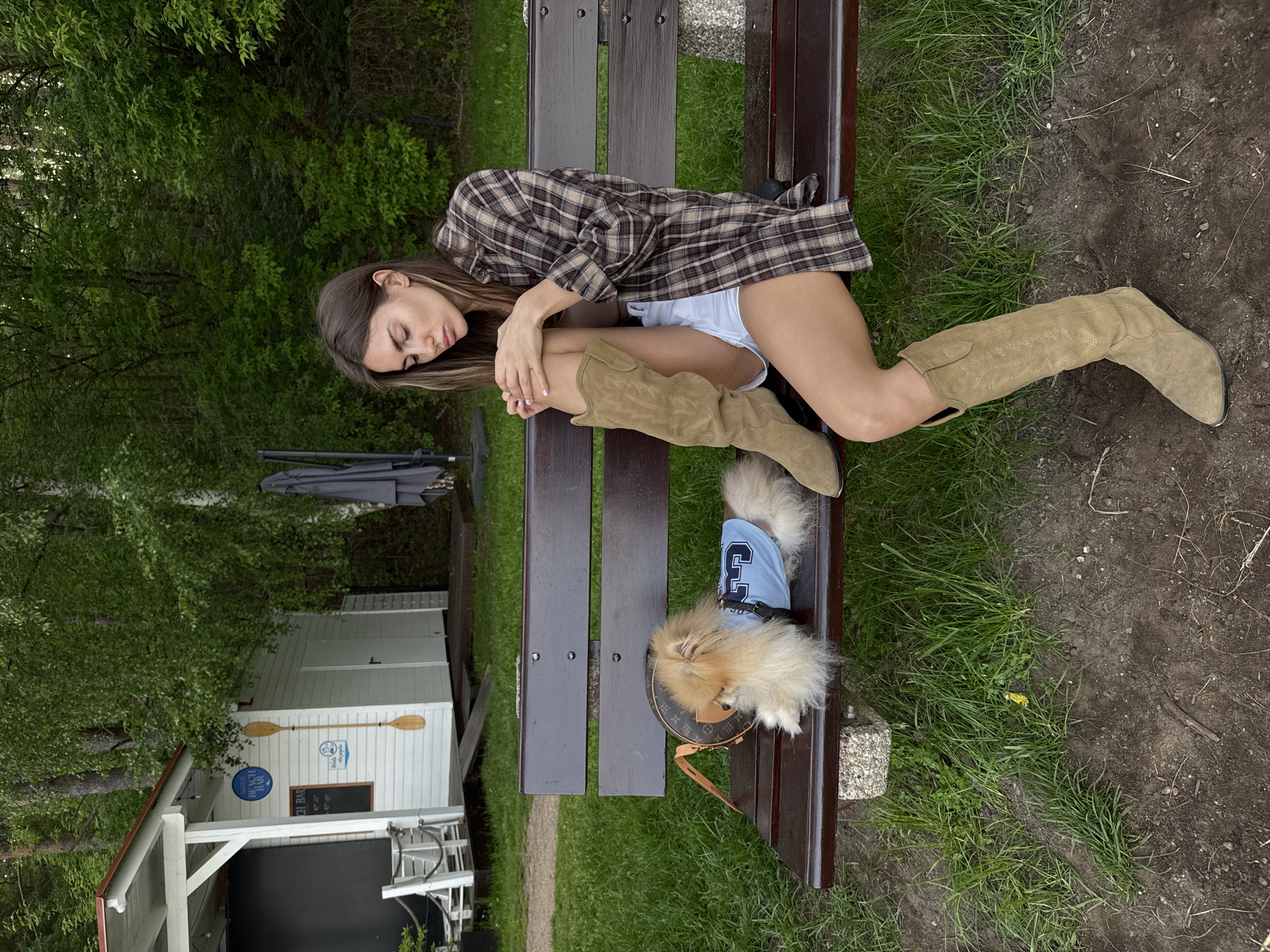 A woman wearing a plaid shirt, white shorts, and knee-high suede boots is lying on a brown wooden bench. Her eyes are closed, and her arms are wrapped around her legs. A fluffy, light-brown dog wearing a blue shirt sits on the bench beside her. Next to the dog is a brown purse. The bench sits on a patch of green grass, and the ground surrounding it is dirt. Behind the bench is a small, white building with oars hanging on the side. The building has a sign that reads "BEACH BAR." Green trees surround the area.