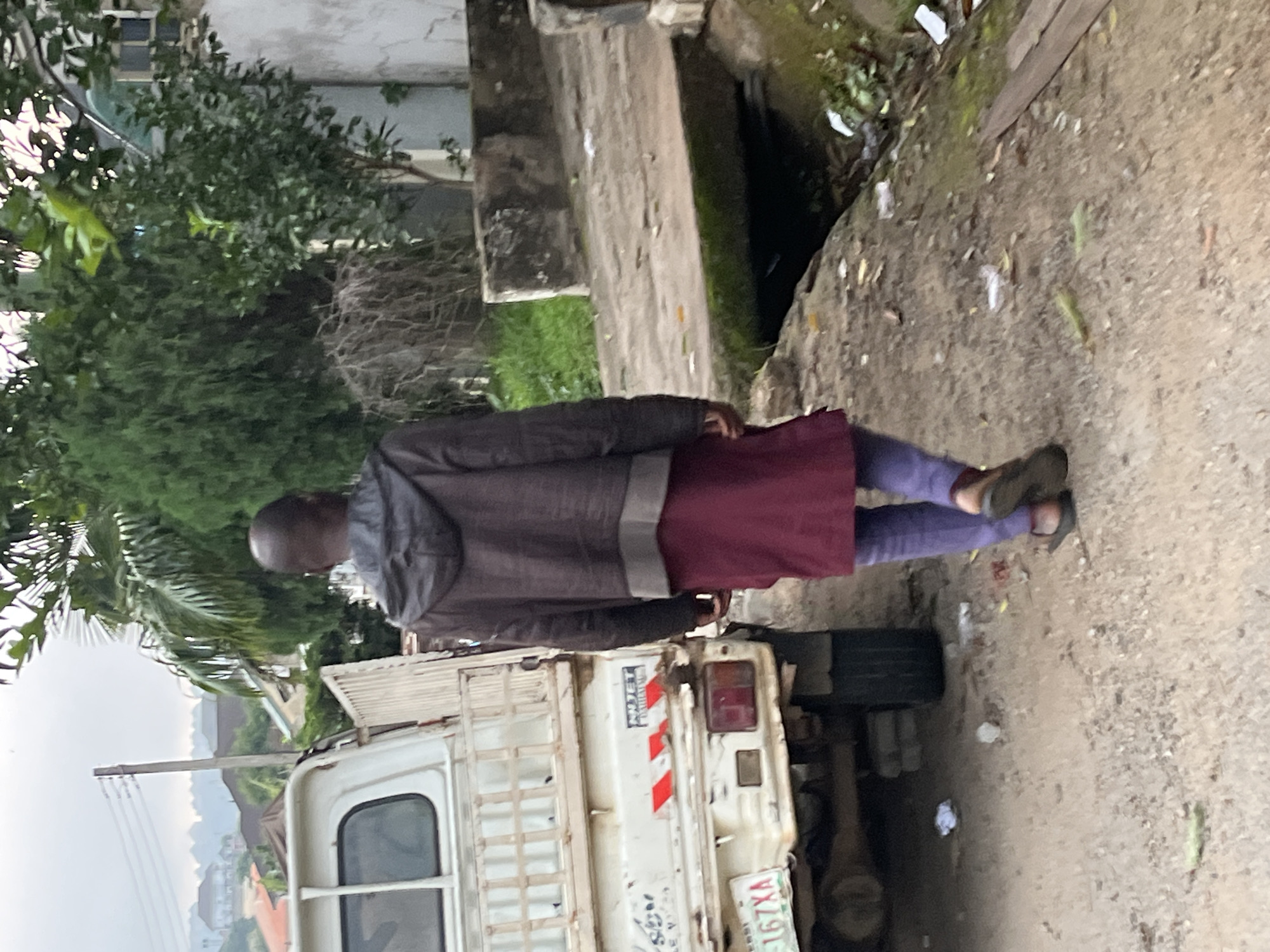 In Abuja, Nigeria, a man with a shaved head walks away from the camera on a dusty path beside a white pickup truck. The man wears a dark hooded jacket over a maroon tunic and light purple pants, with dark sandals on his feet. The pickup truck, seen from its rear passenger side, has a white bed with vertical metal railings and a license plate reading "167XA". Behind the truck, lush green trees and vegetation are visible, along with a concrete structure that appears to be a bridge or elevated walkway over a drainage ditch. The sky is overcast, suggesting a cloudy day. The overall environment appears to be a semi-urban or residential area with a mix of man-made structures and natural greenery.