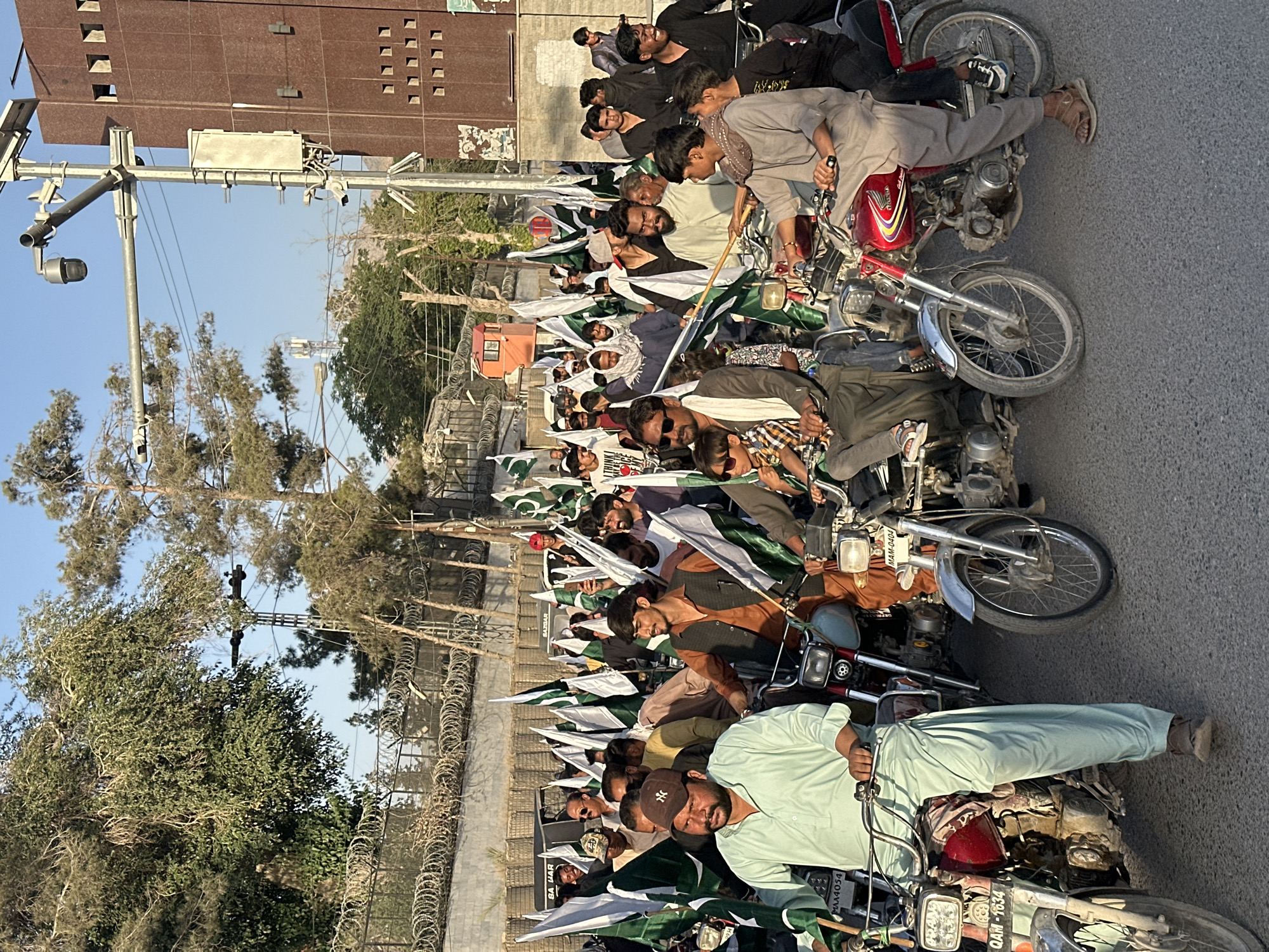 The image depicts a gathering of people on motorcycles in Quetta City Tehsil, Pakistan. The individuals are predominantly male, some wearing traditional clothing and holding Pakistani flags. The motorcycles are parked along a street, lined by a fence with barbed wire. The background features buildings and trees under a clear sky.