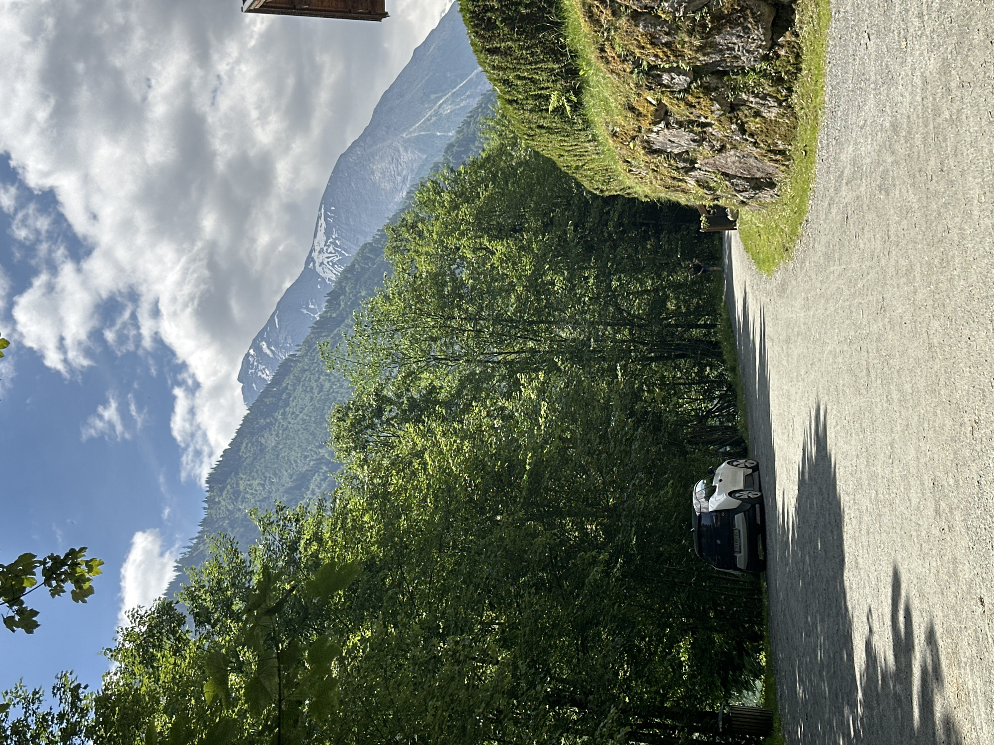 The image shows a scenic view of the Kaprun area in Austria, likely during daytime. A gravel road curves through a lush green landscape. On the left side of the road, a white electric car is parked near a trash can, partially shaded by tall trees. The road is lined with dense green foliage on the left and a moss-covered stone wall with a manicured hedge on the right. In the background, a majestic mountain range dominates the horizon, with its peaks showing patches of snow and its slopes covered in a mix of trees. The sky is partly cloudy, with patches of blue sky visible. A person can be seen walking in the distance on the road. The overall scene suggests a peaceful and natural setting within the Austrian Alps.