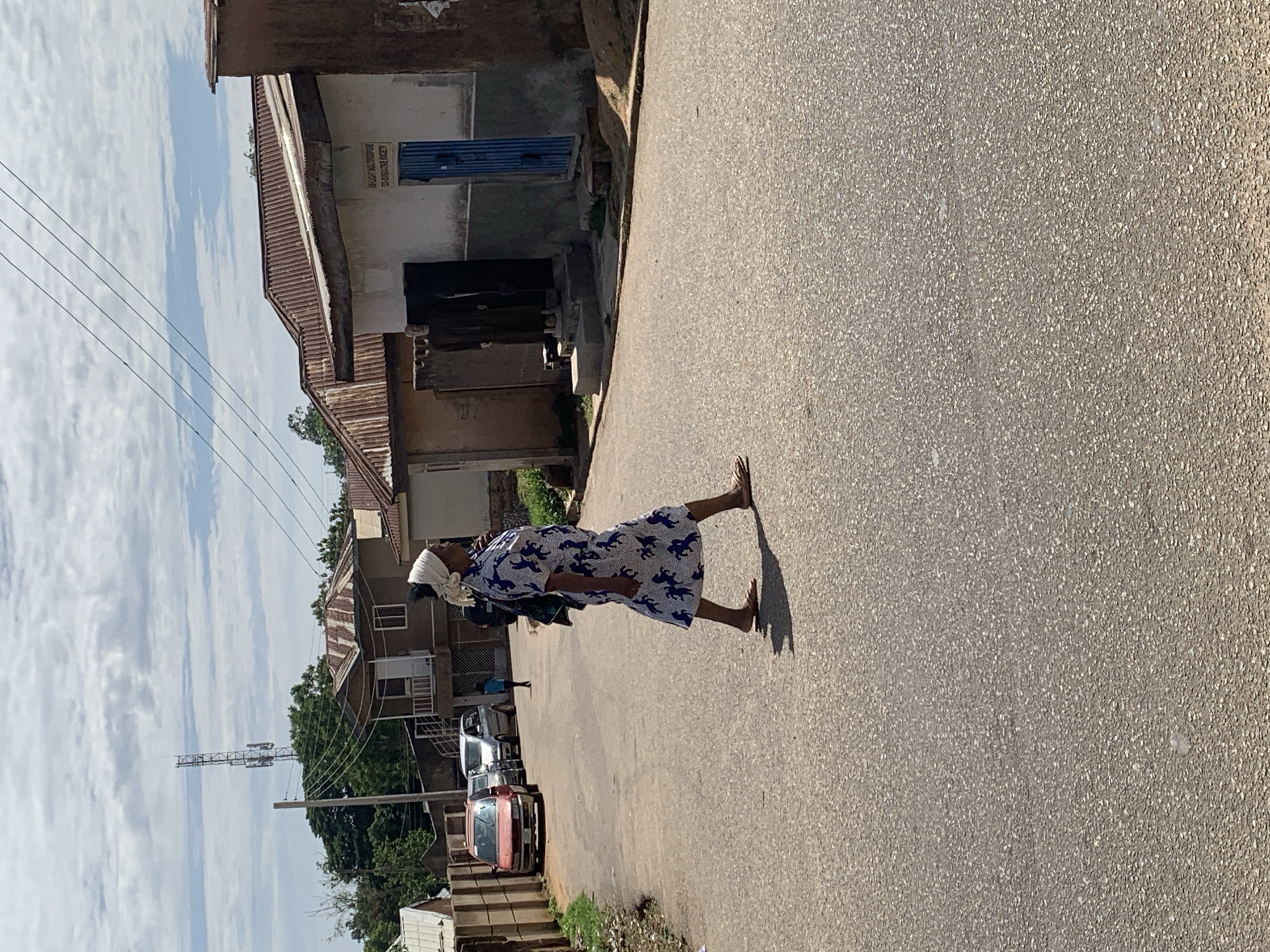 This image depicts a sunny street scene in Jos, Nigeria, characterized by local architecture and a telecommunication mast. A paved asphalt road, showing light-colored aggregate, occupies the foreground and midground.
In the center, a woman with a white headwrap and a blue and white patterned ankle-length dress (featuring a horse motif) walks from left to right, midway across the road. She wears light-colored sandals and carries a dark backpack on her left shoulder. Her shadow is cast diagonally forward to her left, indicating sunlight from the upper right.
On the left side of the road, two cars are parked facing away from the viewer: a red sedan followed by a silver minivan. Further down the road, a small figure in a blue top and dark bottoms walks away from the viewer. A brown perimeter wall borders the left side of the road.
In the background on the left, a two-story building with a brown corrugated metal roof features a white door and a small balcony. Overhead utility poles and wires traverse the upper left quadrant of the image. A tall telecommunication mast with multiple antennas is visible in the far background, rising above dense green foliage under a partly cloudy sky.
On the right side of the image, two structures are prominent. The nearest building has a white and grey facade, a brown corrugated metal roof, and a blue metal door. A sign above the blue door reads "DE-LIGHT MULTIPURPOSE CO-OPERATIVE SOCIETY." A rigid, dark-clad figure, possibly a mannequin, stands in a shadowed doorway adjacent to this sign. Further to the right, another building with a reddish-brown wall is partially visible. The ground on the far right of the road consists of reddish-brown dirt.