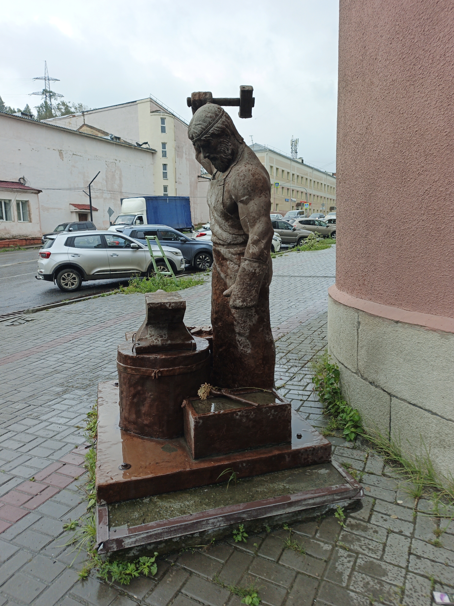 A weathered, rusty metal statue of a blacksmith stands on a wet, paved plaza in Zlatoust, Russia. The blacksmith, depicted with a hammer above his head and leaning forward, is positioned next to an anvil and a trough. The statue is located in front of a salmon-colored building with a stone foundation. In the background, there are several cars parked on a wet street, and a line of buildings with light-colored facades. The sky is overcast. The plaza is made of interlocking pavers, some of which are dark gray and others are reddish-brown. Weeds are growing through the cracks in the paving stones around the base of the statue and the building. The overall scene suggests a slightly damp, possibly cool day.
