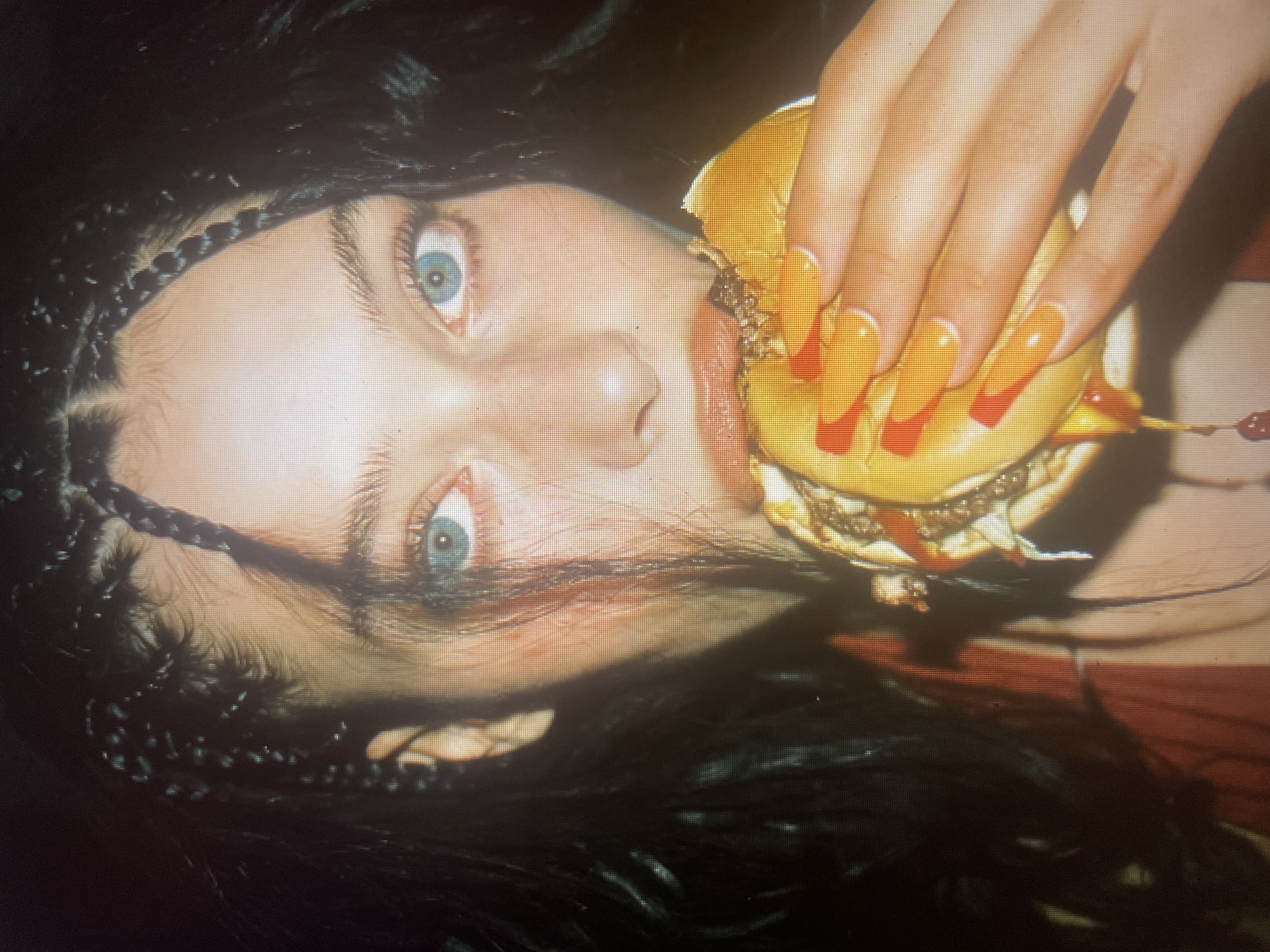 This close-up media file shows a person with striking blue eyes and dark hair styled in braids. The person is holding a hamburger, with their long, orange-tipped nails visible as they grip the food. Ketchup is smeared on the burger and appears to be dripping. The person's face is tilted, and they seem to be in the process of taking a bite. The lighting is dramatic, with a shadow cast across the face, obscuring some features and highlighting others, particularly the eyes. The background is dark and out of focus, with hints of red fabric visible, suggesting a possible setting. The overall impression is one of indulgence and perhaps a touch of edginess, conveyed through the intense gaze and the stylized presentation of the hamburger. There is no visible text in the media file.