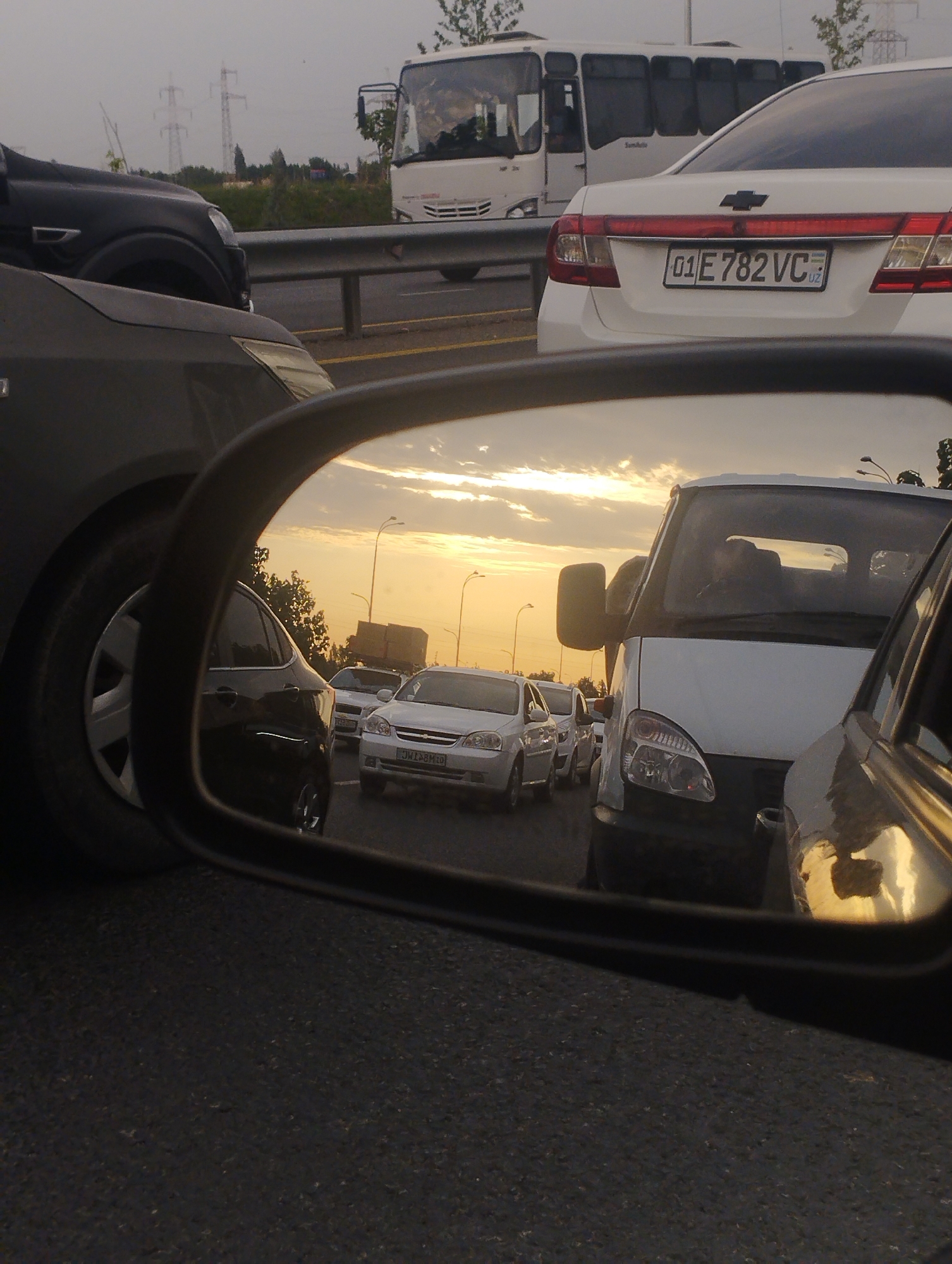 The image is a medium shot of a traffic jam. The foreground is dominated by the side mirror of a car, reflecting a scene of vehicles on a road at sunset. The sky is a hazy orange and yellow, indicating the sun is low on the horizon. Several cars are visible in the reflection, including a white sedan with a license plate that reads "01 E 782 VC UZ". There are also power lines and trees in the background of the reflected scene. In the immediate foreground, parts of two cars are visible: a dark-colored SUV on the left and the side of another vehicle, possibly a white van, on the right. The overall impression is one of congestion and the end of a day. There are no people clearly visible in the image.