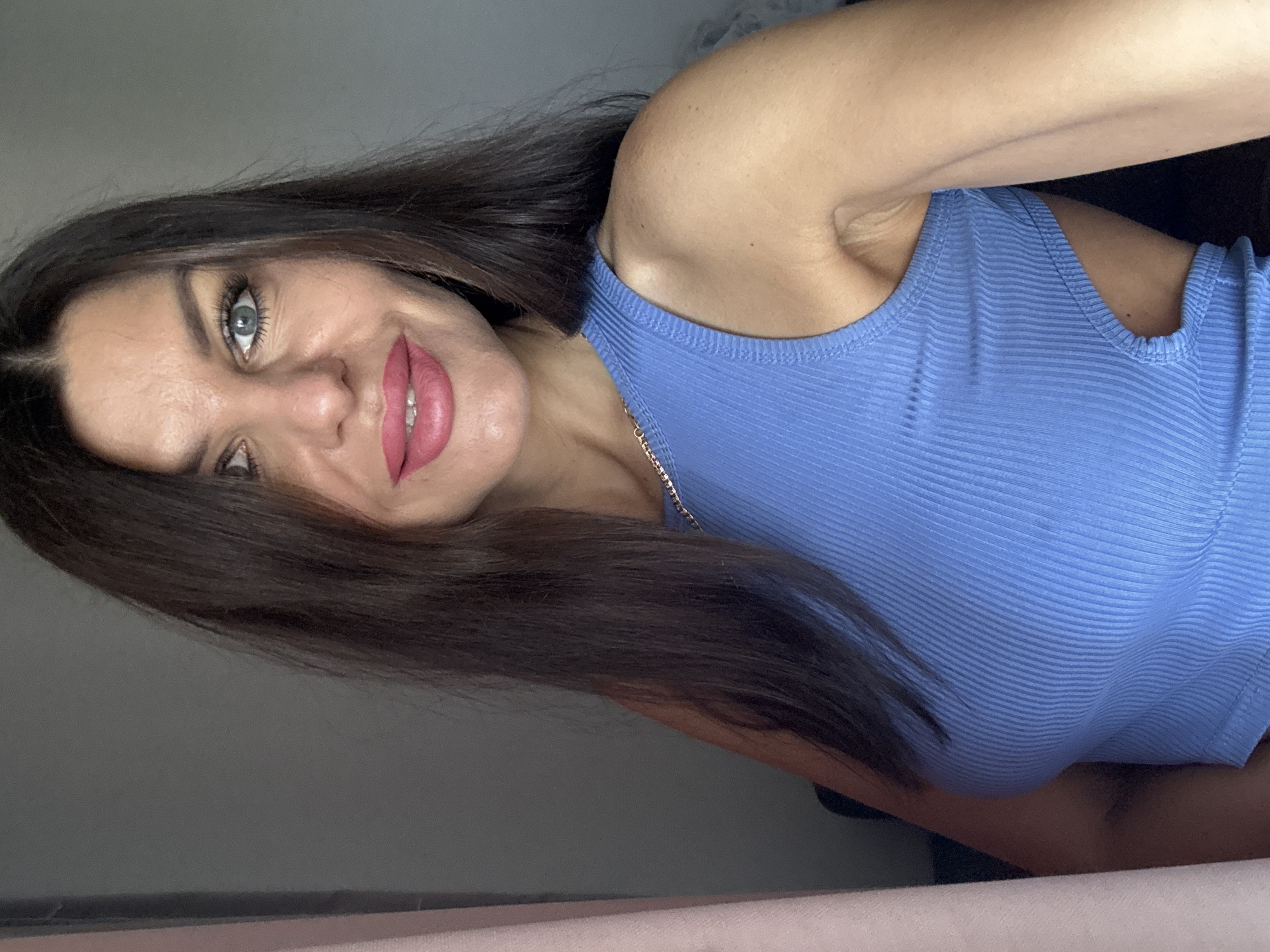 A woman with dark hair and blue eyes, wearing a blue ribbed tank top with a cutout on the side, poses for a selfie. She has on pink lipstick and visible teeth. She is standing indoors, with a plain gray wall behind her. A pink curtain is visible on the left side of the image. The image is taken at a close-up angle, focusing on her face and upper body.