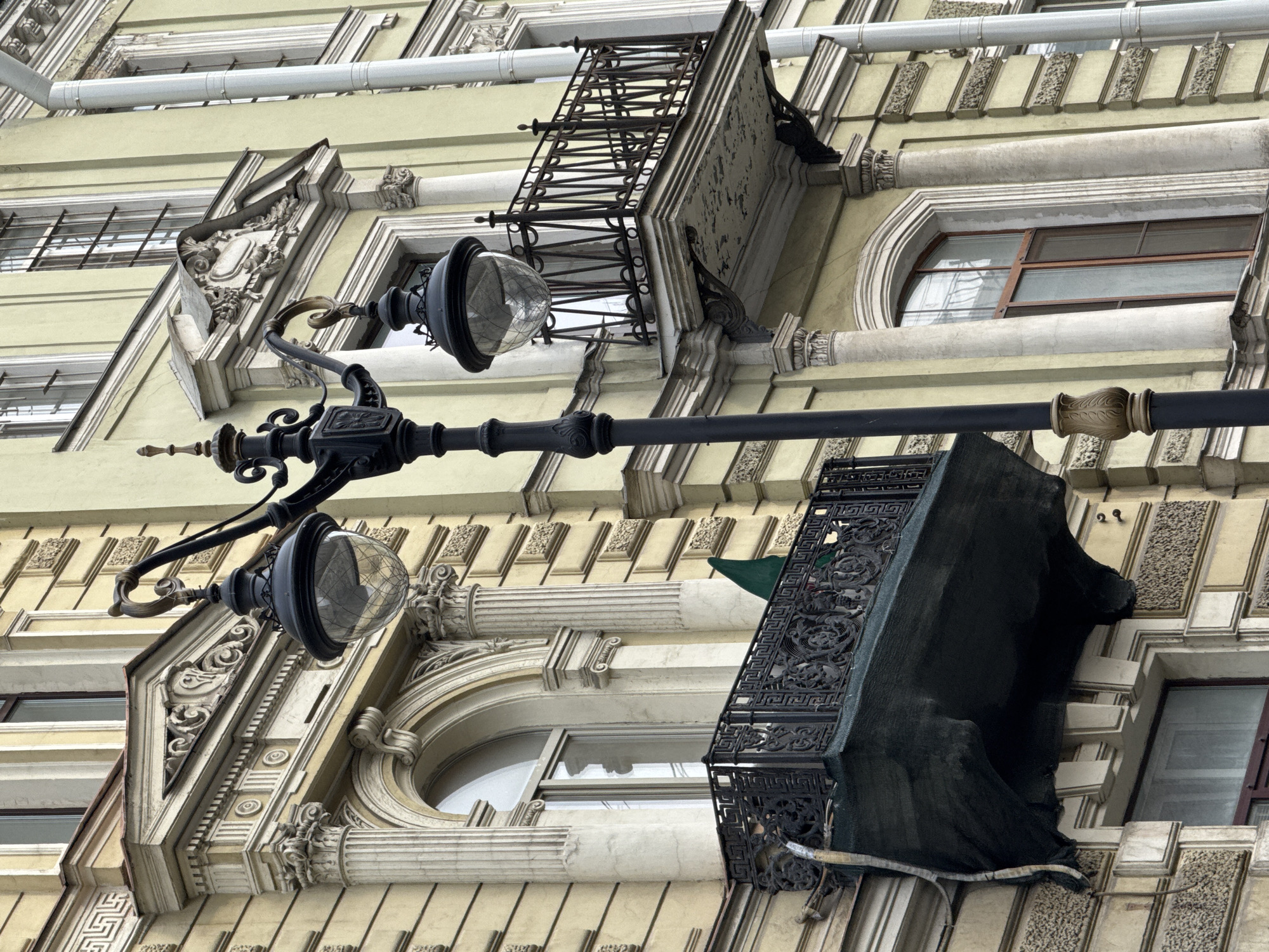 The image depicts the facade of a building in Saint Petersburg, Russia. The building is a pale yellow color and features architectural details such as ornate window frames, columns, and balconies. A black iron street lamp with two globe-shaped lights is mounted on the building. One balcony is covered with a dark material. The style of the building is typical of the city's historic architecture. The angle of the photo is low, looking up at the building.