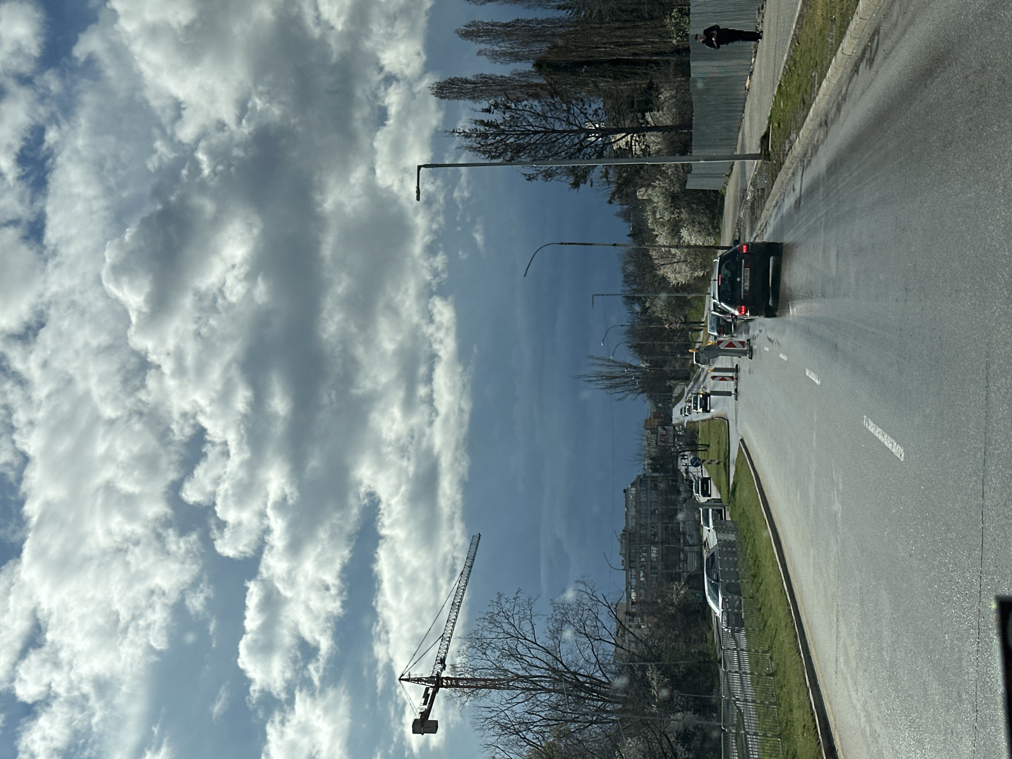 The image depicts a street scene in Sofia, Bulgaria. The view is from inside a vehicle, looking out at traffic on a wet asphalt road. The road has lane markings, and there are several cars, including a dark-colored vehicle in the foreground. Road construction is occurring, indicated by orange and white barriers. Streetlights line the road. A construction crane is visible against a backdrop of cumulus clouds. Buildings, trees, and a metal fence border the road. A person is visible standing on the sidewalk near a fence. The sky is predominantly blue with white clouds.