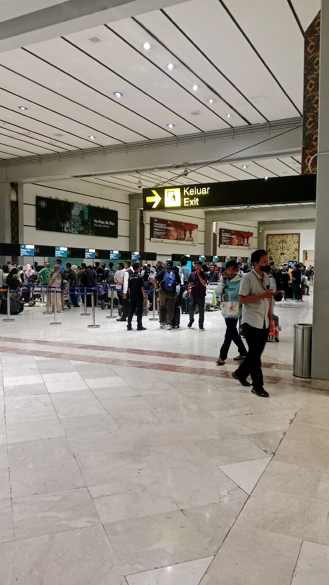 The image depicts an interior view of a busy airport terminal, specifically at the Soekarno-Hatta International Airport in Tangerang, Indonesia. The scene is characterized by a high ceiling with white panels and black linear accents, illuminated by recessed lighting. A prominent yellow and black sign with an arrow pointing to the right reads "Keluar Exit," indicating an exit route.
In the foreground, a man wearing a light-colored shirt and black pants walks towards the right, his attention seemingly directed away from the camera. His left hand is raised, and he holds a dark object. The floor is made of polished marble tiles with red and white inlay patterns.
Further back, a queue of people is visible at a series of check-in or baggage drop counters, labeled with numbers 60, 61, 62, and 63. Many individuals are standing with luggage, some are interacting with airline staff or at the counters, while others are walking through the terminal. Several large advertisements are mounted on the walls behind the counters. The overall environment suggests a typical, active airport setting.