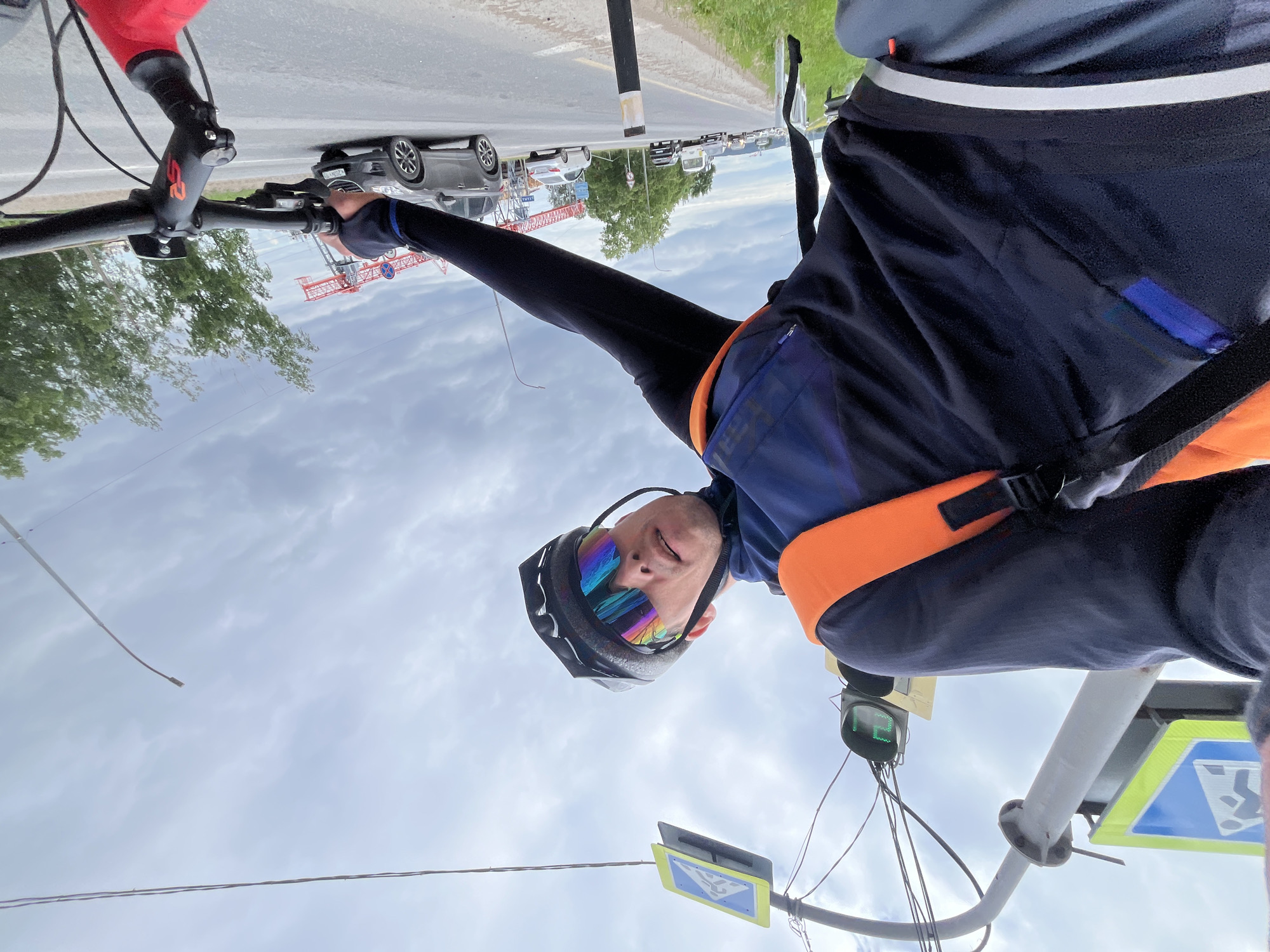 A first-person perspective video still captures a cyclist in Chaikovsky, Russia, riding on a road adjacent to a pedestrian crossing and traffic light. The cyclist, wearing a black helmet and multicolored reflective sunglasses, is looking forward at the camera. They are dressed in a dark blue cycling jacket with an orange vest worn over it. The cyclist's left hand is visible on the handlebars of a bicycle, which is red in color. The road is paved and has a white line marking the edge of the lane. Several cars, a bus, and a white sedan are driving on the road. In the background, trees line the roadside, and a construction crane can be seen. The sky is overcast with clouds.