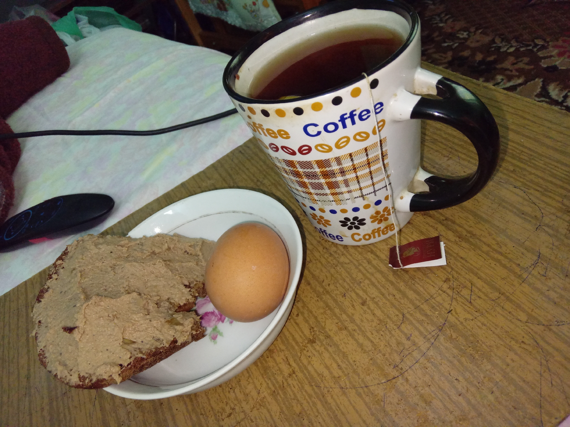 A breakfast scene unfolds on a wooden table. A white mug with a black handle, adorned with colorful polka dots and the word "Coffee" repeated, is filled with tea. A tea bag string dangles from the mug, with a small red tag attached. To the left, a white plate holds a slice of dark bread generously spread with a beige-colored pate. Adjacent to the bread is a single, light brown, hard-boiled egg. In the background, a rumpled white cloth is visible, with a dark red, knitted item to its left. A black electronic device, possibly a remote control, rests on the cloth. The wooden table shows signs of wear, with scratches and markings. The overall impression is of a simple, home-cooked breakfast. The image is set in Kitskany, Moldova.