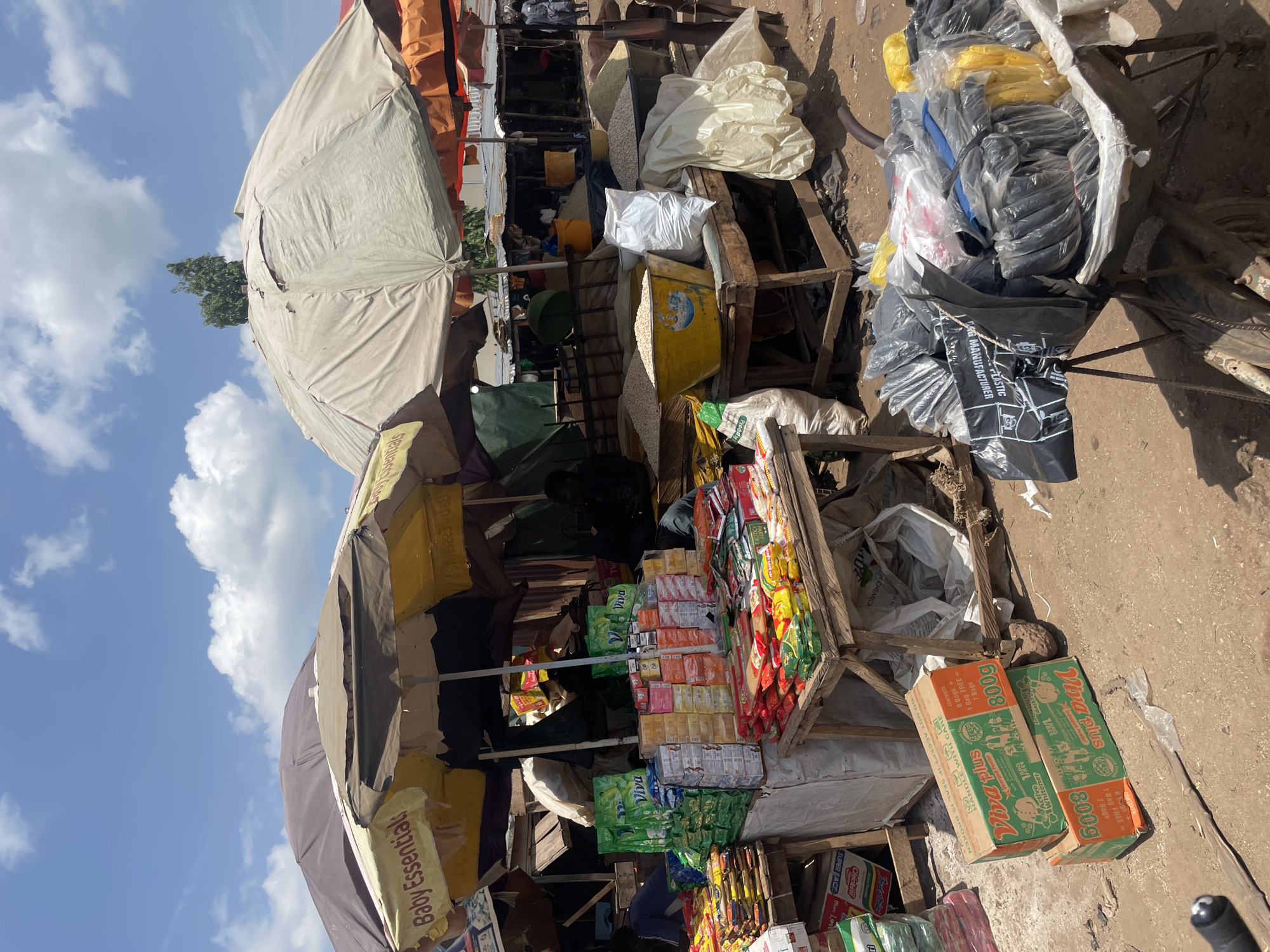 Here's a detailed description of the image:
**Scene:** The image depicts a bustling marketplace in an urban area.
**Environment:** The ground is dirt or compacted soil. Various stalls are set up, covered by umbrellas and makeshift awnings, against a blue sky with scattered clouds.
**Objects:**
* Various goods are on display, including boxed detergents, snack foods, and packaged items stacked on wooden stands. Some of the brands/products are labeled with names such as "Viva Plus," "Baby Essentials", and "SUPER FACE ML."
* Large bags of materials are stacked behind the displays.
* Plastic bags, both black and yellow, are seen near the top right of the frame.
**People:** Several people are seen moving around and interacting within the market stalls, including shopkeepers and customers.
**Interactions:** The scene suggests active buying and selling of goods within a crowded marketplace.