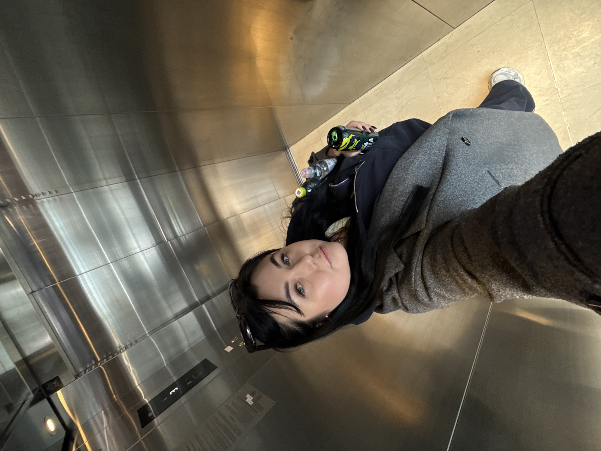 A young woman is captured in a selfie-style photograph, taken from a slightly upward and rotated angle, inside what appears to be an elevator. She has long, dark hair and light-colored eyes, looking directly at the camera with a neutral expression. Sunglasses are resting on top of her head. She is dressed in a dark blue hooded garment layered under a grey, textured coat. Her left hand, adorned with dark nail polish, holds a green and black branded energy drink can. Several other plastic bottles, including one with a yellow cap, are visible on the floor next to her.
The elevator interior features polished, reflective metal walls that show shadows and bright reflections of light sources. The floor is covered with light-colored, possibly beige or cream-colored, tiles. A white sneaker is visible on the floor, suggesting another person or her own foot in the frame. On the lower left side of the metal wall, there's an elevator control panel with the illuminated number "3" prominently displayed, indicating the current floor. Below this, there's a small rectangular display with some indistinct symbols and text. A partially visible sign with unreadable text is also present on the wall near the bottom left corner. The lighting appears to be artificial, typical of an indoor environment. The overall impression is a candid moment captured within a modern building in Perm, Russia.