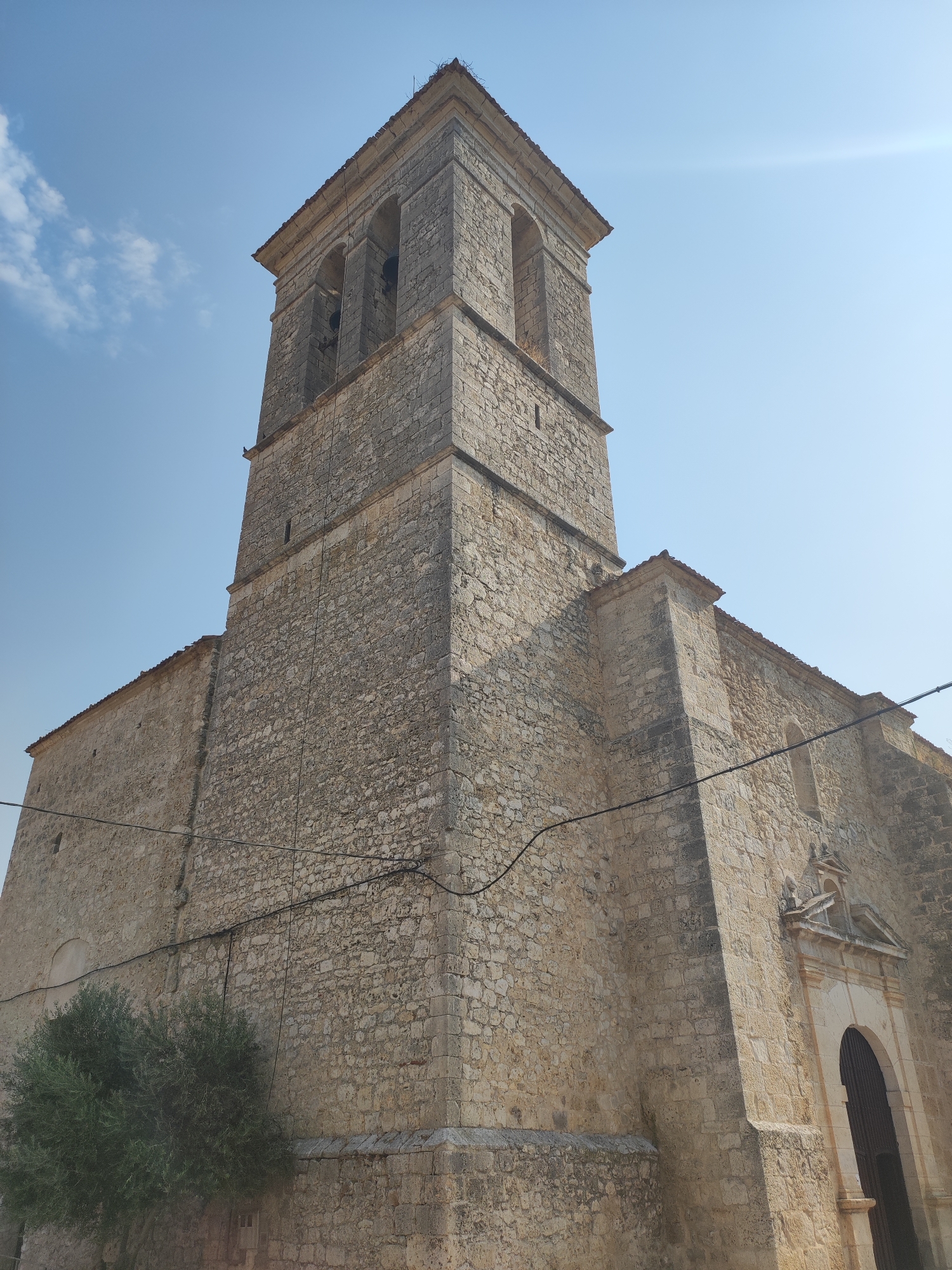 The image shows a stone church with a bell tower in Traspinedo, Spain. The church is made of rough-hewn, light-colored stones. The bell tower is square and has arched openings with bells visible inside. A red-tiled roof runs along the main body of the church, and the bell tower has a flat top with a cornice. A dark, arched wooden doorway is visible on the right side of the church. To the left of the church, a green tree with dense foliage is present. The sky is clear and blue with a few wispy white clouds. Electrical wires run across the building. The perspective is low-angle, looking up at the church.
