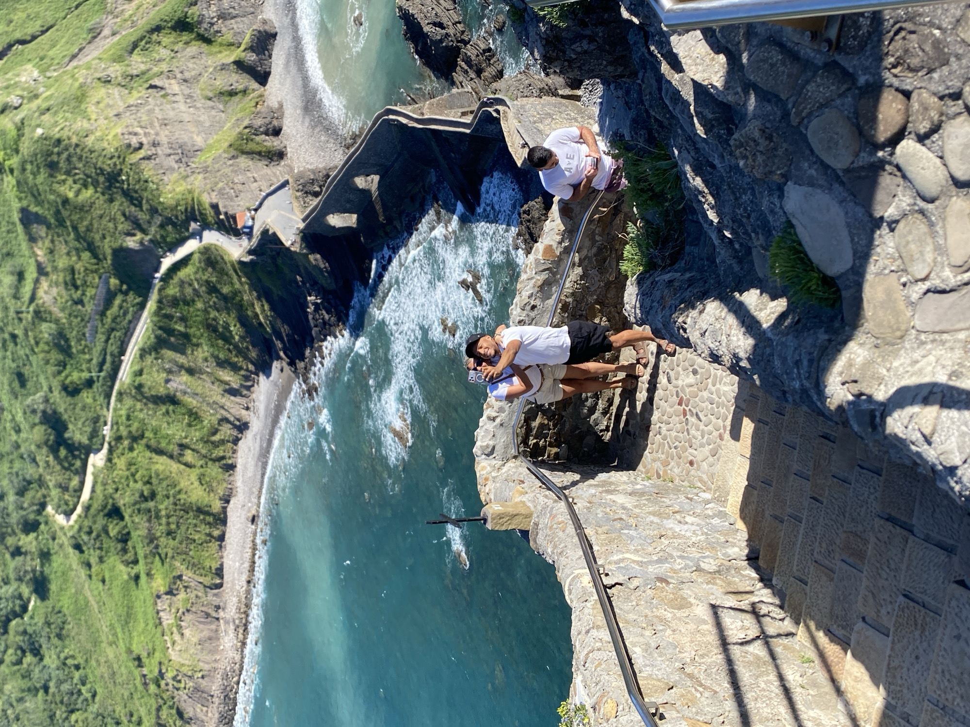 The image depicts a scenic coastal view at San Juan de Gaztelugatxe, near Bakio, Spain. In the foreground, a stone staircase descends, flanked by stone walls. Three individuals are visible on a paved landing. Two people, a man and a woman, stand together taking a selfie. The man wears a white t-shirt and black shorts, and the woman wears a white t-shirt and beige shorts. Both are wearing sandals. Another man is standing further down the stairs, looking at his phone. He wears a white t-shirt with "AVE" printed on it and maroon shorts. The background reveals a dramatic coastline with a rugged, green-covered cliff face. A winding road with cars is visible traversing the cliff. A narrow, stone bridge with arched openings leads to a small island topped with a structure, likely a hermitage. The turquoise ocean waves crash against the rocky shore. The scene is bathed in bright sunlight, casting distinct shadows.