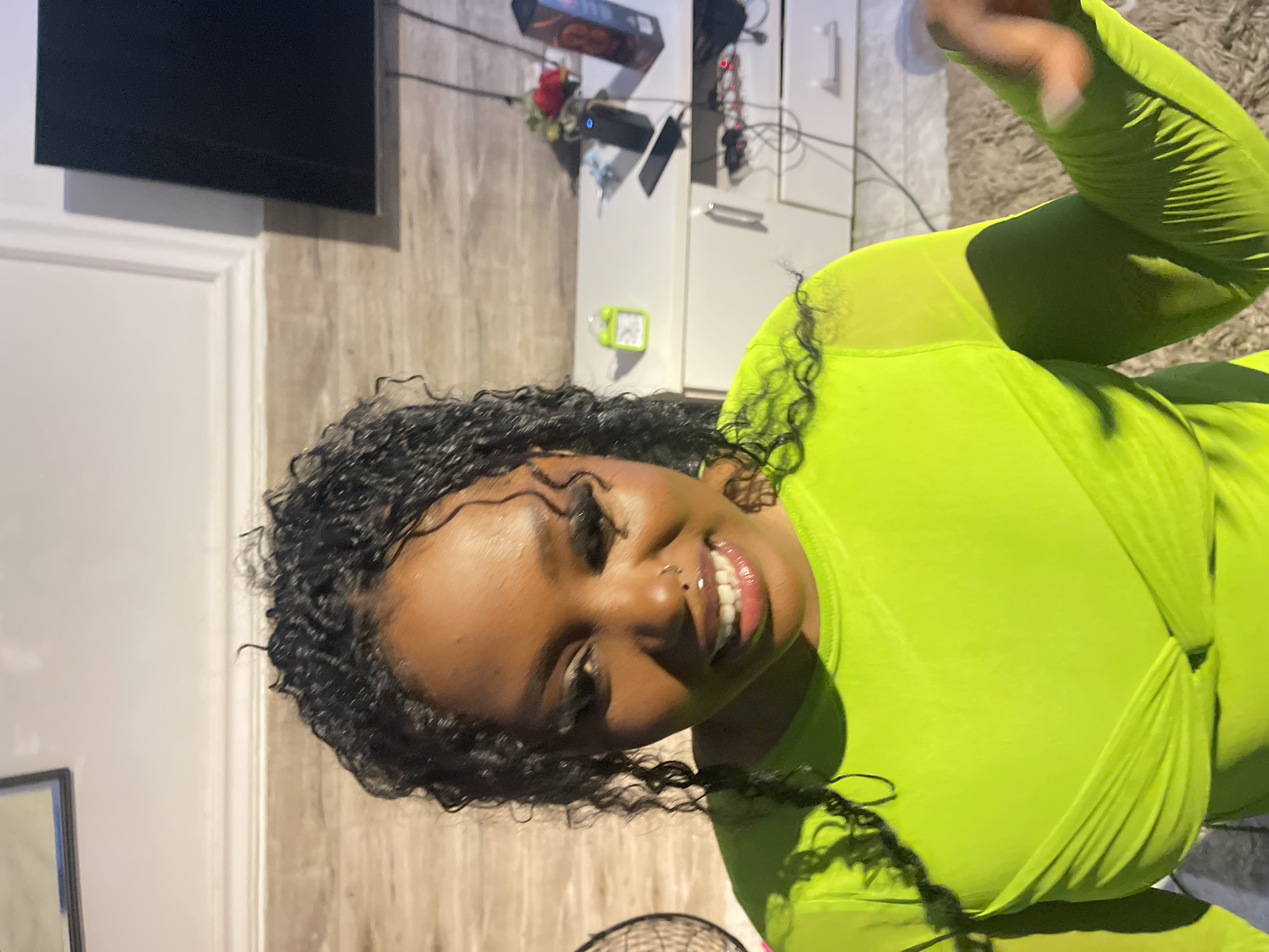 A vibrant portrait-style image captures a young Black woman smiling broadly indoors, likely in a residential setting in Kuchingoro, Nigeria. The woman is positioned centrally, looking directly at the camera with a cheerful expression, showcasing a wide smile and a nose stud piercing. She has dark, curly hair and is wearing a striking neon green, long-sleeved top or jumpsuit made of a lightweight, possibly sheer, fabric. The garment features soft gathering around the chest.
The background reveals a domestic environment. To her left, a light-colored wall with a subtle wood-like texture is visible, along with a white door frame. A black flat-screen television is mounted on this wall. To her right, behind her, a white cabinet or dresser holds several items, including a dark box, a red rose, various wires, and what appears to be a power strip. A small, bright green digital device, possibly an alarm clock or timer, hangs on the cabinet. Part of a light-colored, textured rug or carpet is visible on the far right, and the grille of a fan can be seen at the bottom of the frame. The bright illumination highlights the woman's face, contributing to the overall cheerful mood of the photograph. No discernible text is visible on any objects within the image.