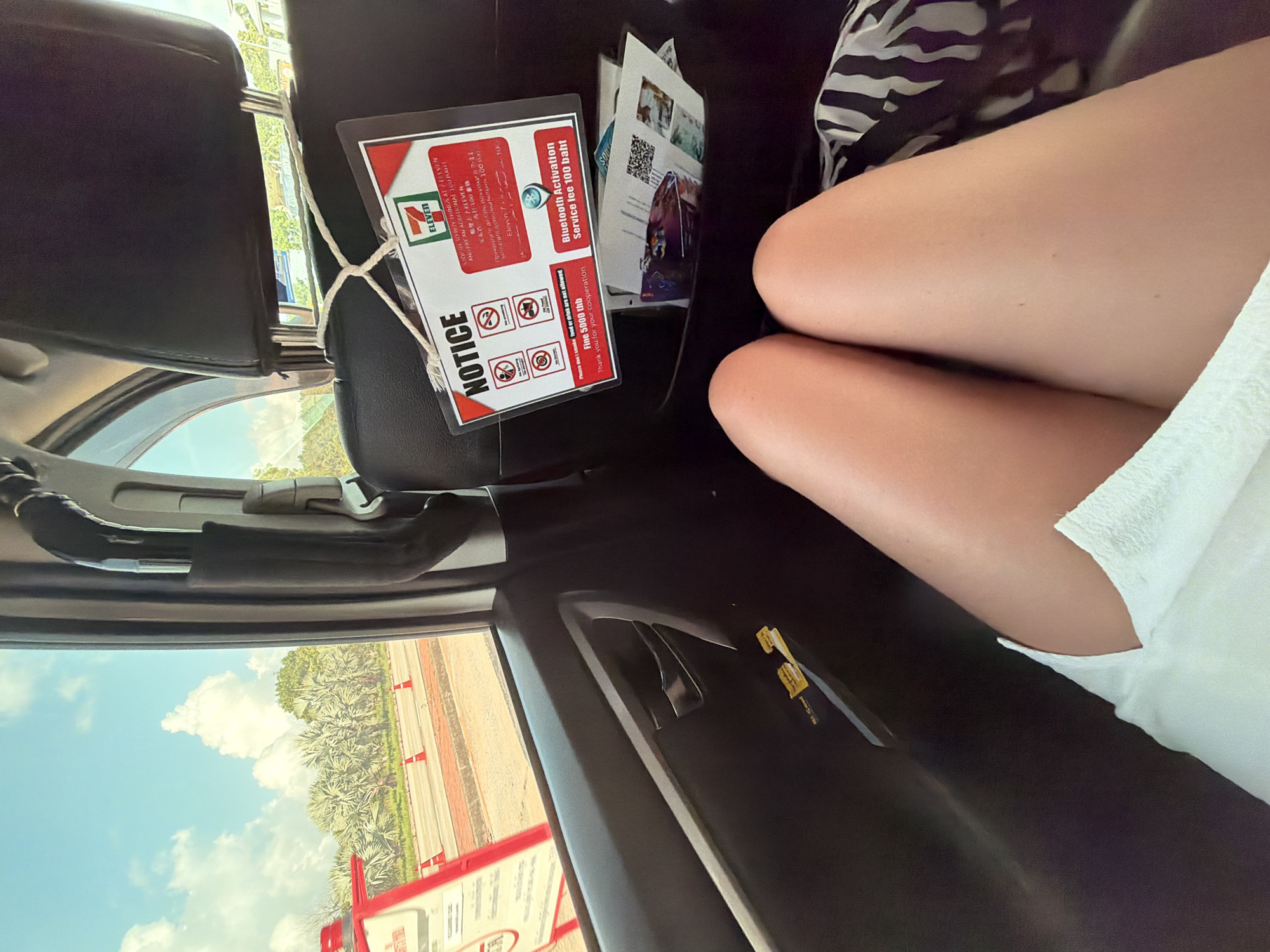 The image shows the interior of a vehicle, likely a taxi, on a sunny day. The view is from the backseat, looking towards the front. In the foreground, a person's legs, clad in a white garment and a black and white striped fabric, are visible.
A notice from 7-Eleven, a convenience store chain, is hanging from the headrest of the driver's seat. The notice, written in English and possibly other languages, displays several prohibition symbols (e.g., no smoking, no eating/drinking, no littering). It also mentions a "Bluetooth Activation Service fee 100 baht."
Outside the car window, a road is visible with traffic cones and lush green vegetation, including palm trees. The sky is blue with scattered white clouds. The environment suggests a location in a tropical or subtropical region.
Given the reference to 7-Eleven and the visual cues of tropical flora, the location is likely in Thailand. While the image itself does not explicitly state the location as "บ้านท่าฉัตรไชย, Thailand," it is consistent with such a setting. The scene depicts a moment of travel, possibly waiting in traffic or at a stop.