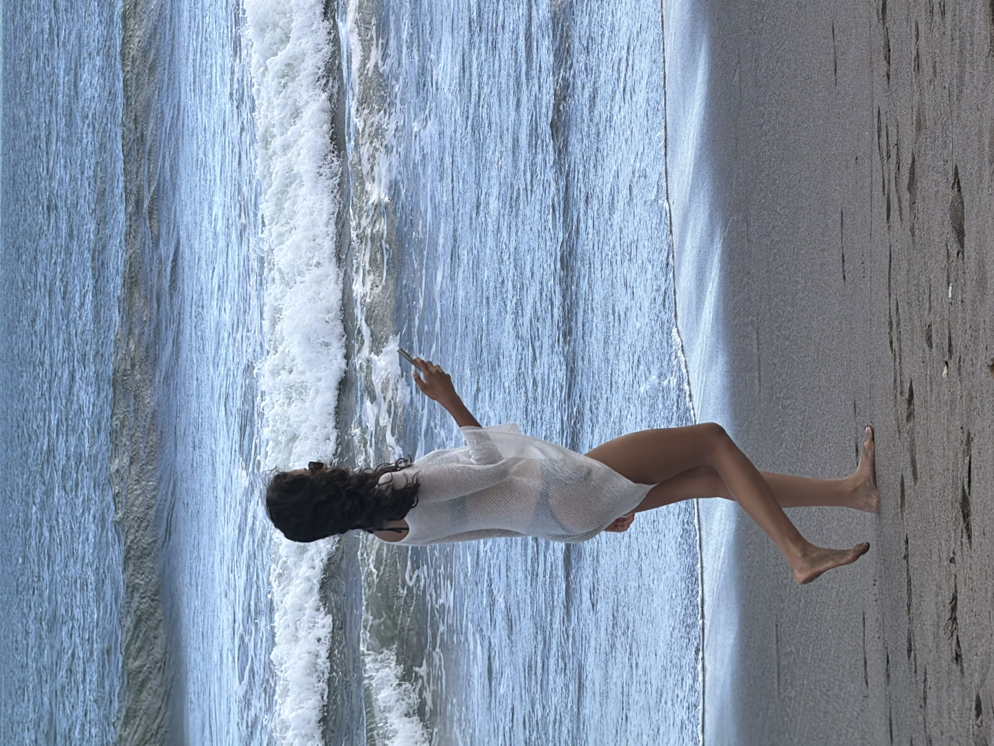 A woman walks on a beach in North Kuta, Indonesia, near the ocean. She is wearing a white cover-up over a bikini and sunglasses. She is holding a smartphone in her right hand. The ocean is blue with white waves, and the sand is dark. The scene is captured during the day.