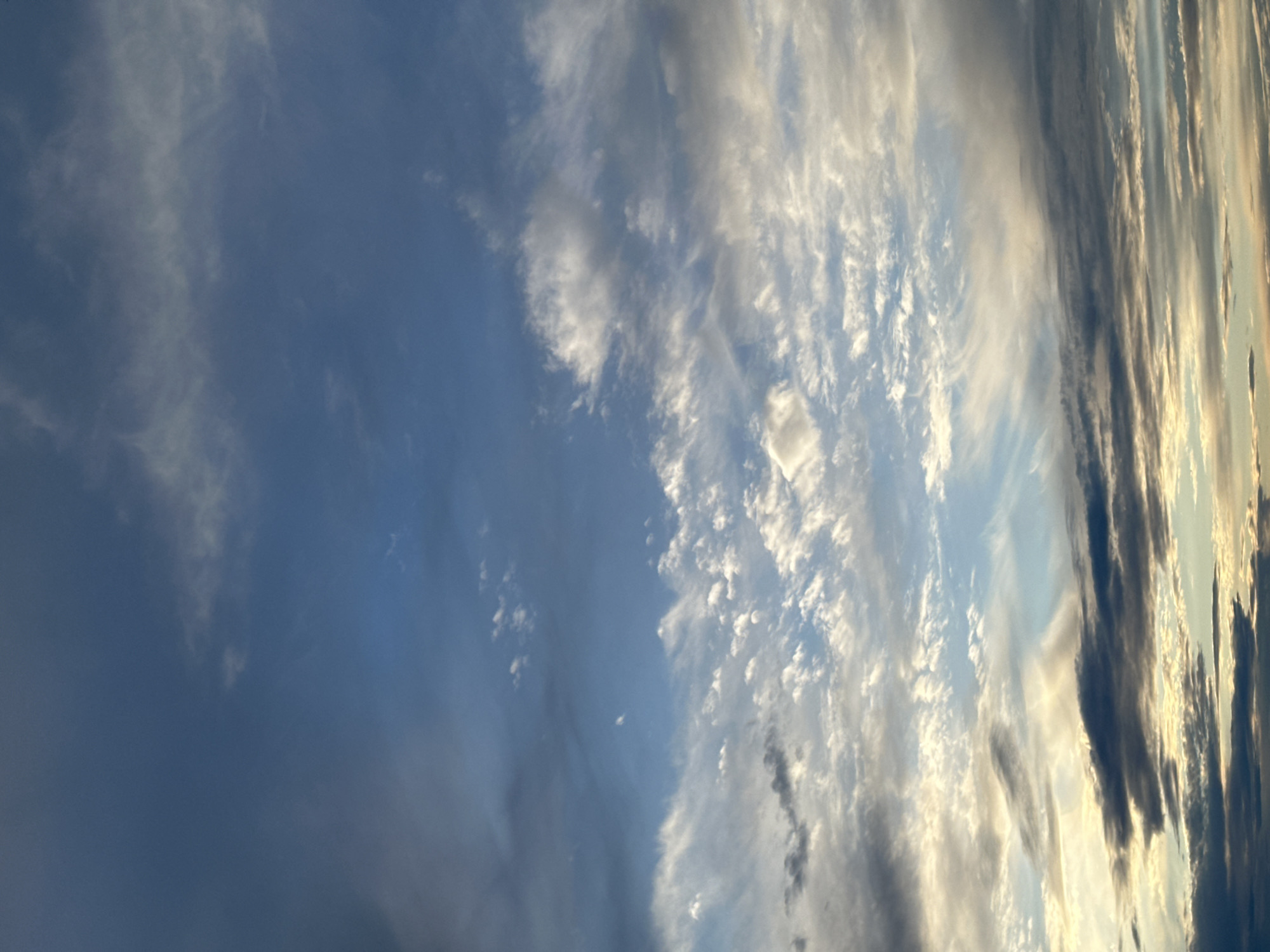 The image depicts a sky filled with various types of clouds. On the left side, there are darker, denser clouds. The central area features small, puffy clouds. To the right, long, striated clouds are present. The sky's color ranges from a deep blue to lighter, brighter tones, indicating varying degrees of sunlight. The location is indeterminate from this image.