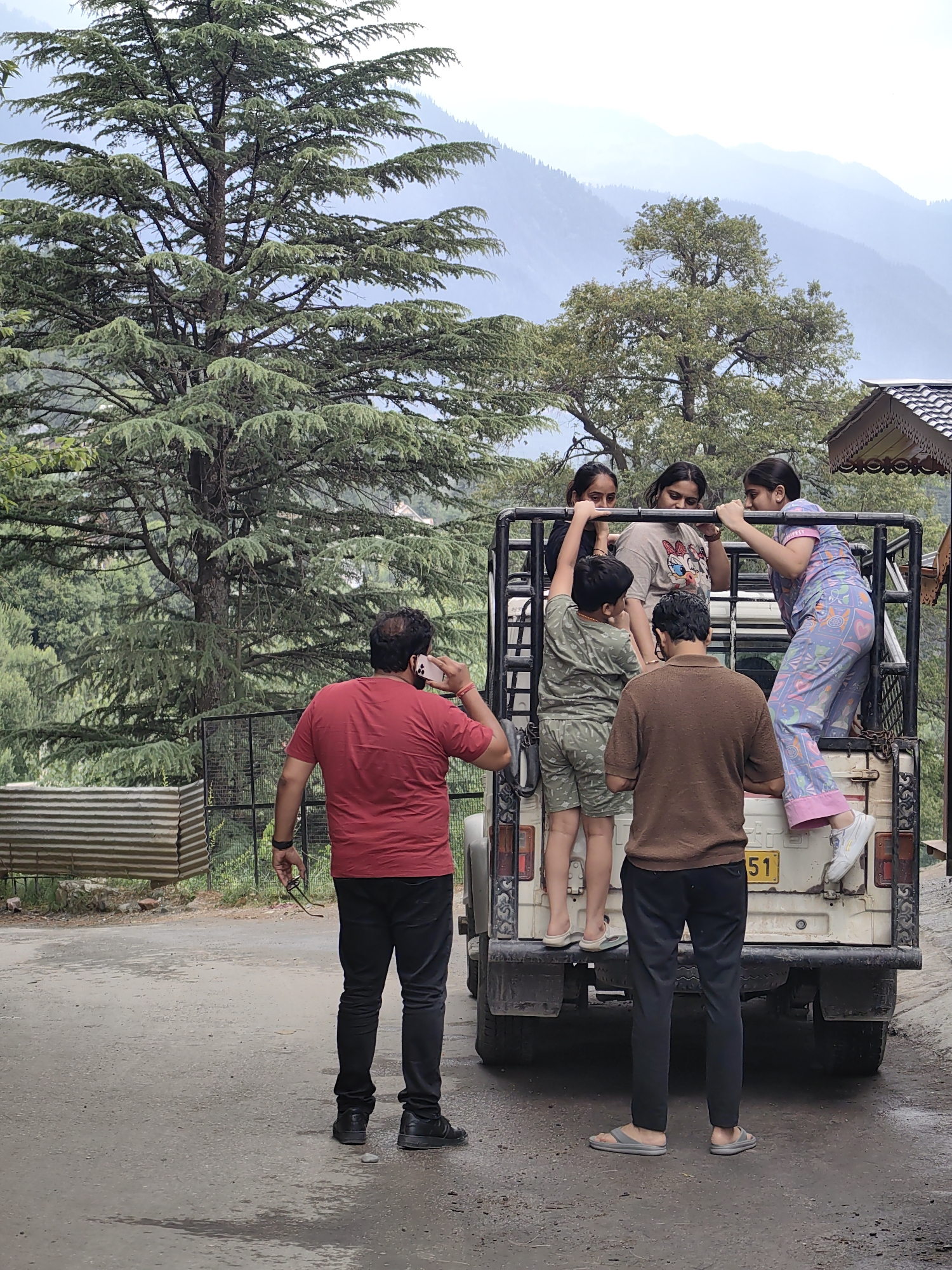 In Vashisht, India, four young people, three girls and one boy, are positioned inside and on the back of a white, open-top jeep. The boy, wearing a green camouflage-patterned shirt and shorts, stands within the vehicle. One girl, in a pink and blue patterned pajama set, is leaning against the rear of the jeep with her legs over the tailgate. Another girl, in a gray t-shirt with a cartoon graphic, stands beside her. A third girl, wearing a black top, is visible behind them. Two men are on the ground. One man, in a red t-shirt and black pants, is standing and holding a phone to his ear. The second man, in a brown shirt and black pants, stands facing the rear of the jeep. The scene is set against a backdrop of lush green trees and rolling hills, with a cloudy sky. A corrugated metal structure is visible to the left of the jeep.