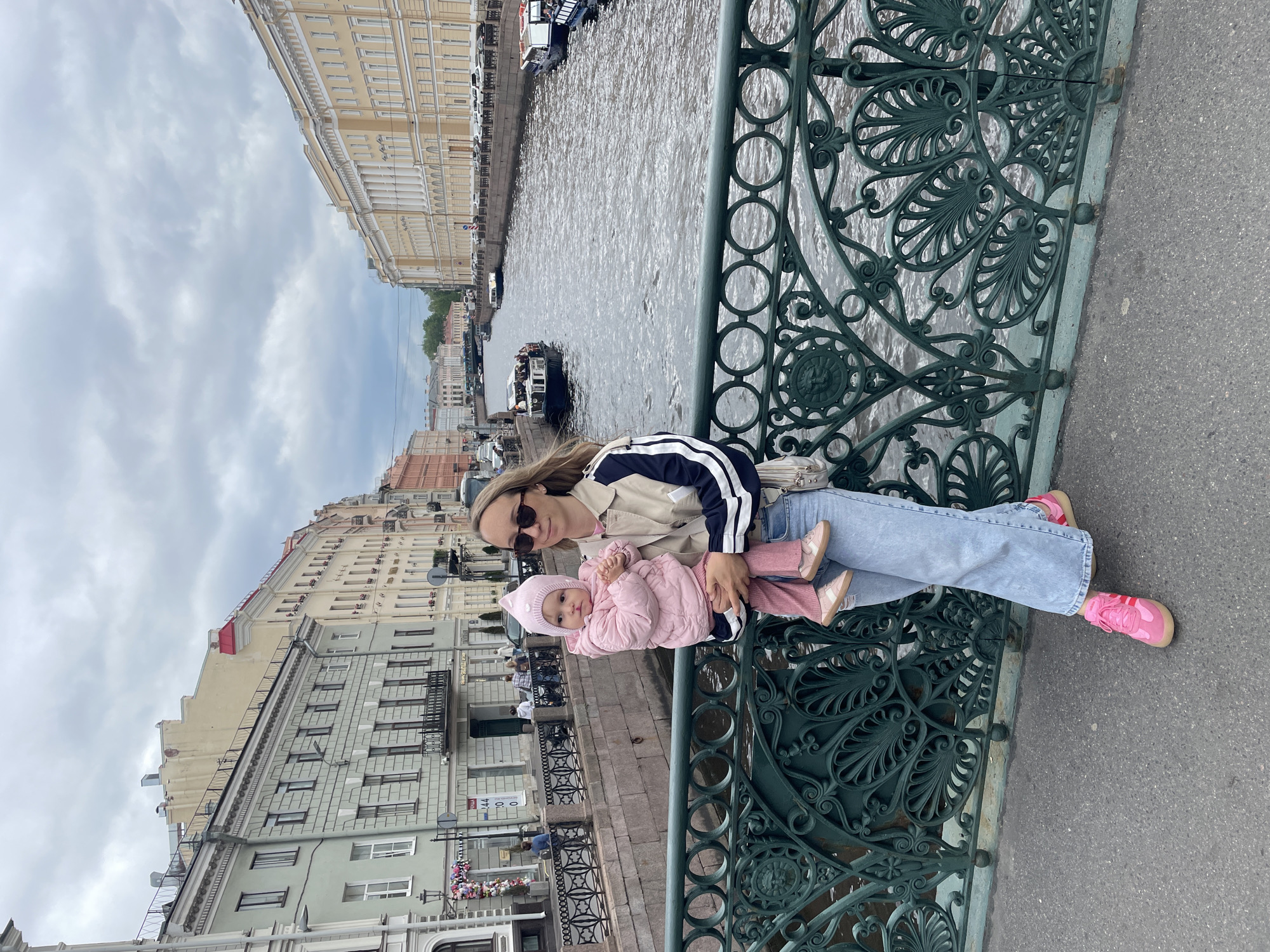 A medium-shot captures a woman and a baby seated on the decorative green metal railing of a bridge, with a canal and buildings in the background. The woman, sporting sunglasses, wears a beige jacket with dark stripes, light blue jeans, and bright pink sneakers. She is holding the baby, who is clad in a light pink padded coat, a matching pink hat, and light pink shoes.
The architectural backdrop reveals the cityscape of Saint Petersburg, Russia. The canal runs between buildings of various heights and architectural styles, including one with a prominent yellow facade. Small boats can be seen on the water. The sky is overcast with cloudy conditions. The bridge's railing features intricate metalwork with a repeating pattern of leaf-like motifs and circular details. A portion of the bridge's grey asphalt surface is visible on the right side of the frame.
