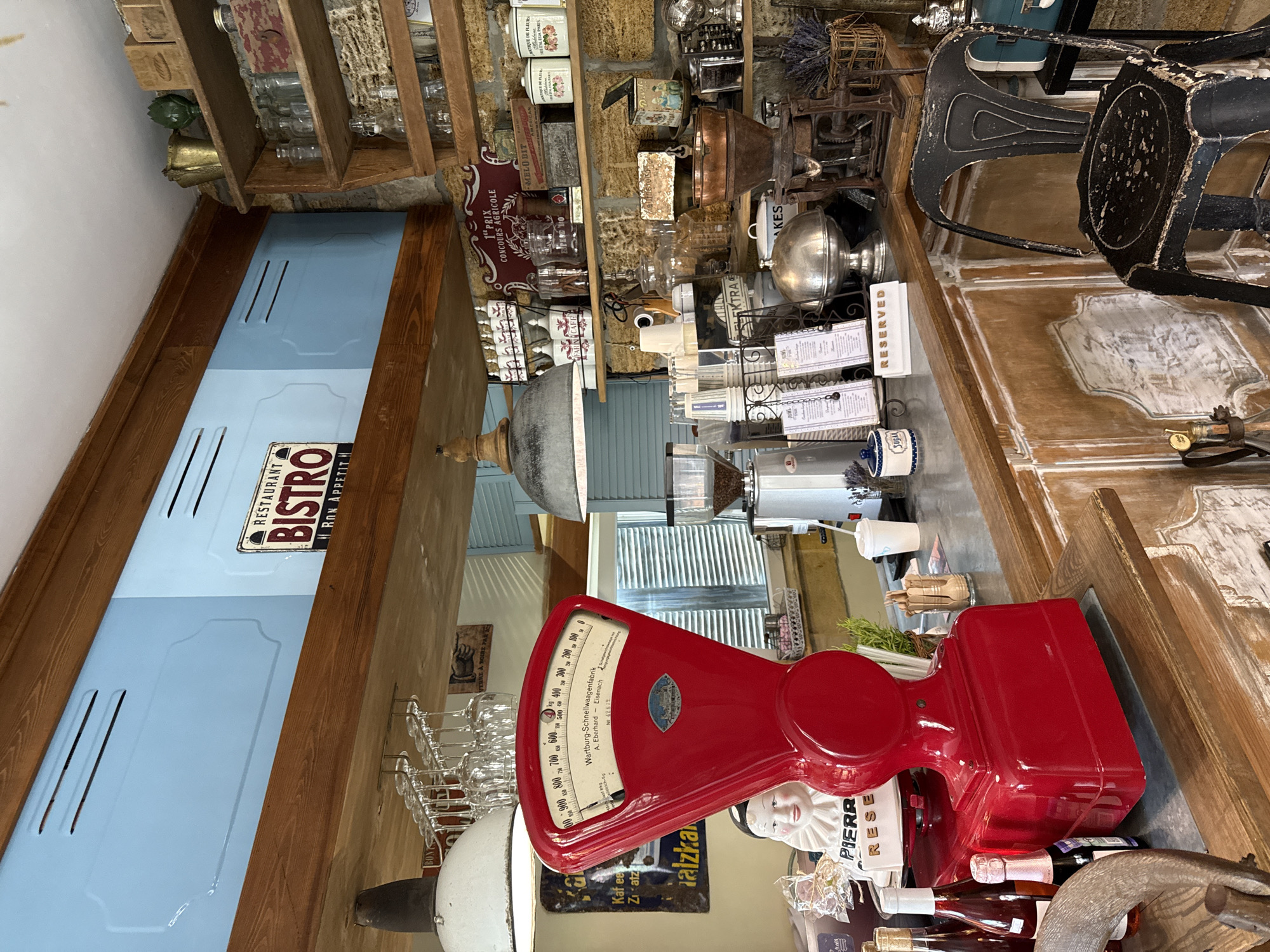 The image shows the interior of a restaurant or cafe with a rustic and vintage aesthetic. The foreground is dominated by a bright red vintage scale and several bottles, some appearing to be wine bottles. To the left of the scale, a porcelain doll with a ruffled collar is visible. Behind the scale, a counter is set up with a coffee grinder, cups, and various other items.
The background reveals shelves filled with jars, bottles, and decorative items, including what appears to be dried lavender. A sign reading "RESTAURANT BISTRO" with "H. Ron Appetit" underneath is mounted on a wooden beam. The walls are a pale blue with decorative insets, and the ceiling beams are made of dark wood. A black metal chair is positioned in the lower right corner of the image, suggesting a seating area. The overall impression is one of a charming, old-fashioned establishment. The location is the city of Natukhaevskaya, Russia.