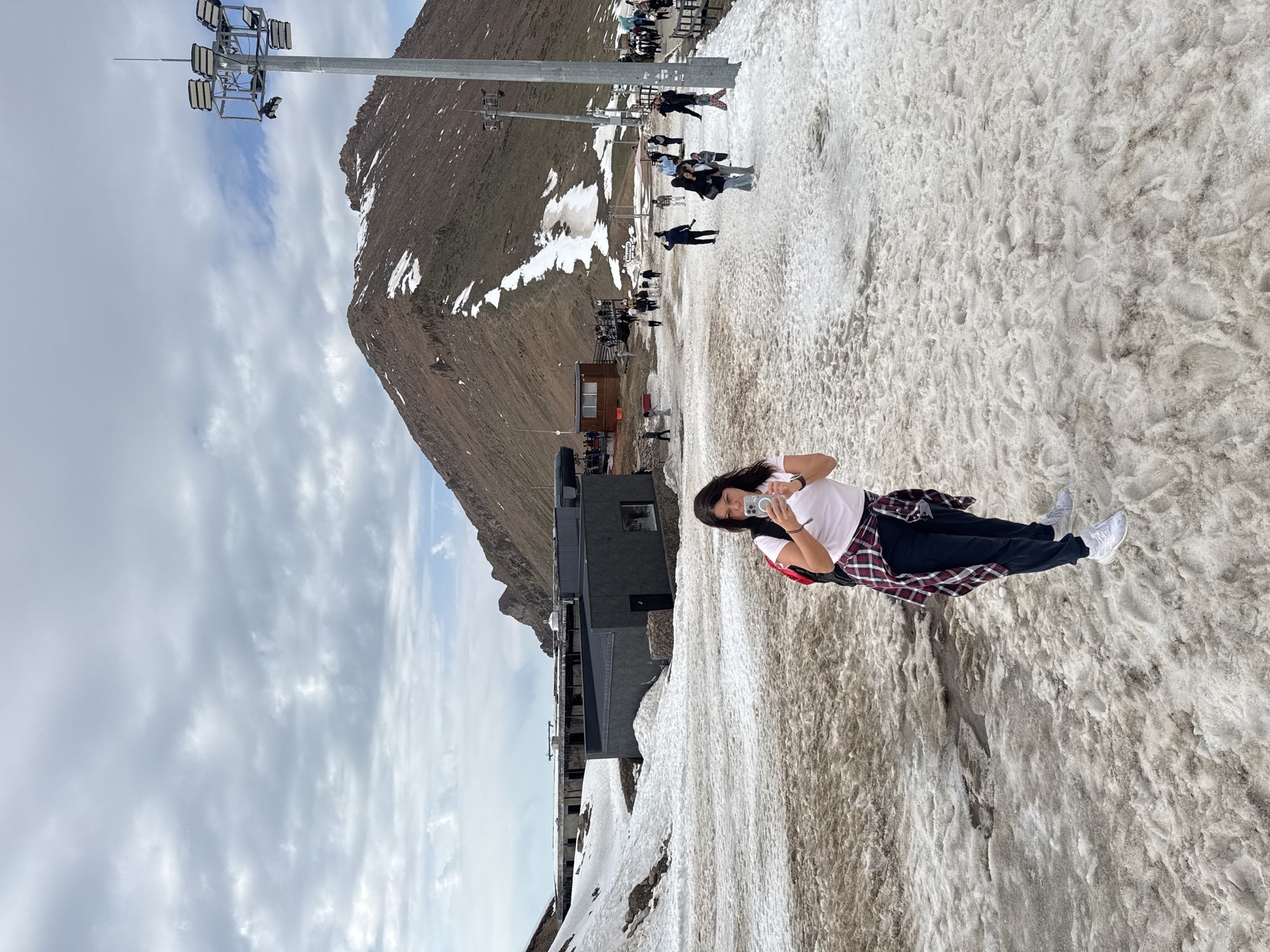 A woman is lying on a snow-covered surface taking a photo with her phone. She is wearing a white t-shirt, dark pants, white shoes, and has a red and black flannel shirt tied around her waist. Behind her is a mountainous landscape with a large building and a ski lift. In the background, other people are visible. The location is likely in the mountains near Almaty, Kazakhstan, known for its ski resorts and mountainous terrain.