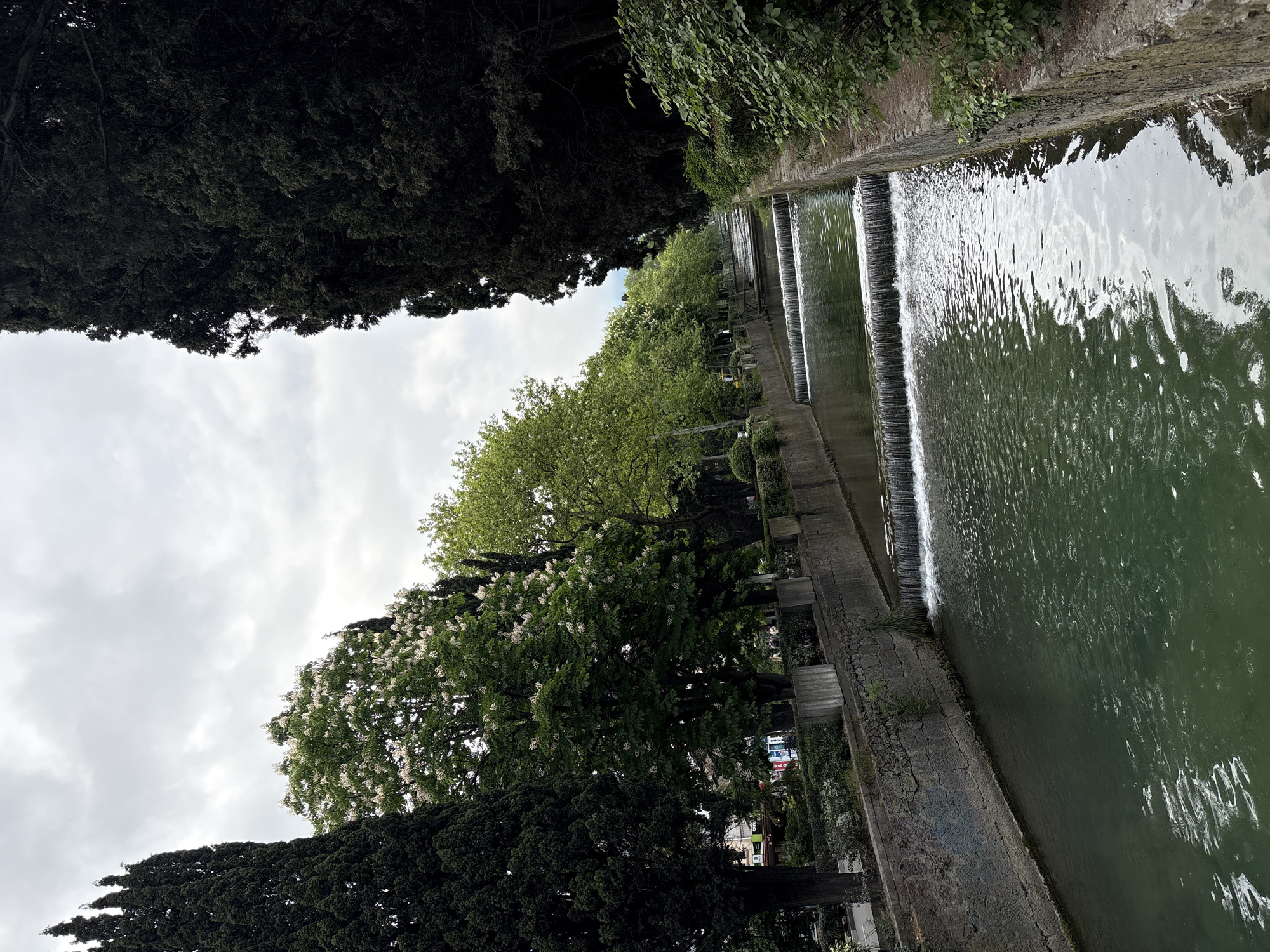 A water canal with small cascading waterfalls runs through a city. Lush greenery, including tall, dark trees and blooming trees, line the sides of the canal. The sky is cloudy. The location is город Ялта, Ukraine.