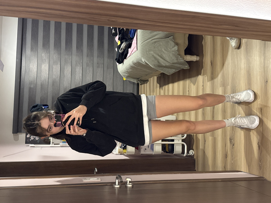 A person stands in front of a mirror, taking a selfie. They are wearing a black hoodie, gray shorts with white trim, and white sneakers. The reflection shows a bed with gray bedding, clothes piled on top, and a roller shade on the wall. The floor is light wood, and a bathroom door with a chrome handle and faucet is visible below the mirror. There is also a small white rolling cart in the background.