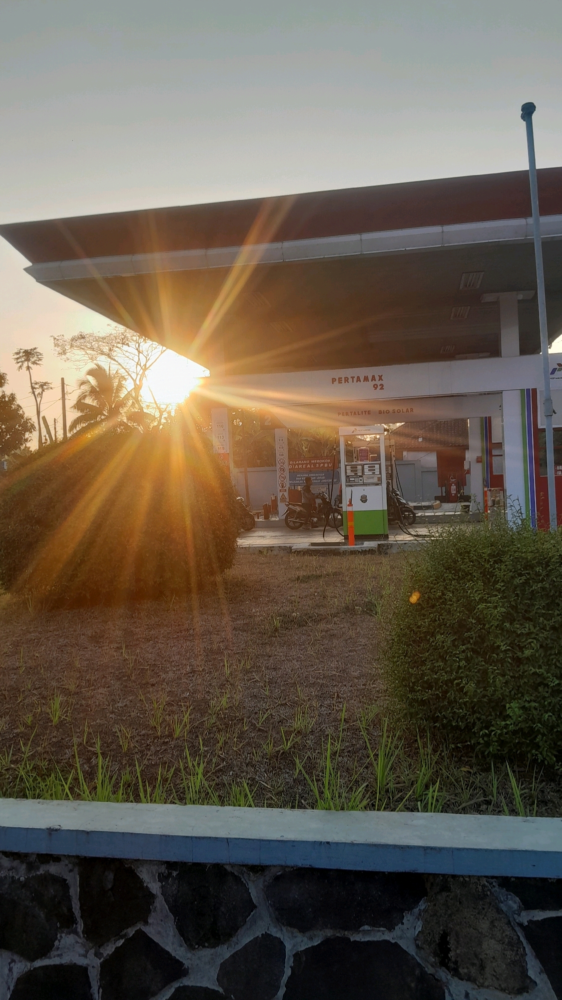 The image depicts a gas station, identified as Pertamina, with the brand "PERTAMAX 92" prominently displayed. The sun is setting, casting a warm, golden glow and creating visible light rays that emanate from behind a large, rounded bush in the foreground. The gas station features multiple pumps, one of which is colored green and white, and the signage also indicates "PERTALITE BIO SOLAR." Two motorcycles are parked near the pumps. The ground in front of the gas station appears to be covered in dry grass and dirt, with a stone wall and a concrete border visible in the lower part of the frame. In the background, there are trees and buildings, suggesting an outdoor setting likely within a developed area. The presence of the Pertamina brand and the types of fuel advertised strongly suggest this is a gas station located in Indonesia, specifically in the city of Subang, as indicated.