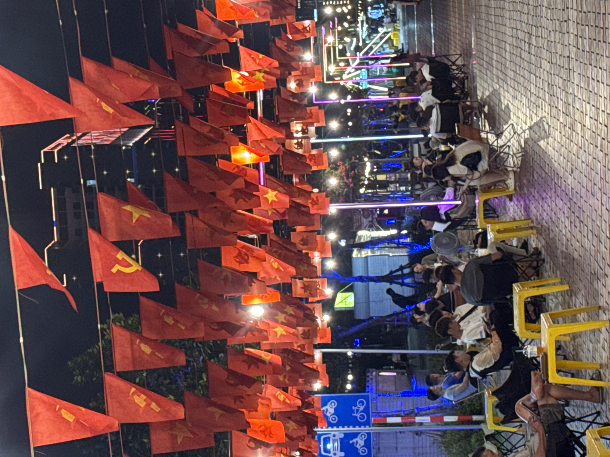 The image captures a vibrant nighttime scene in an outdoor urban setting, likely a street food area or outdoor café in Đà Nẵng, Vietnam. Numerous people, mostly young adults, are gathered, seated on small yellow plastic chairs around low tables, socializing, eating, and drinking.
Overhead, a dense canopy of red flags is strung across the space, brightly illuminated by string lights. The majority of these flags are the national flag of Vietnam, featuring a yellow five-pointed star on a red background. Interspersed among them are flags displaying a yellow hammer and sickle symbol, representing the Communist Party of Vietnam.
The background is filled with various artificial lights, including vertical neon lights in shades of blue, pink, and purple, contributing to a lively, festive ambiance. The ground beneath the tables is paved with light-colored interlocking tiles. In the lower right corner, blue road signs with white pictograms for motorcycles and bicycles are visible, indicating proximity to a road or parking zone. The time of day is clearly night, as evidenced by the dark sky and the reliance on artificial lighting. The atmosphere appears casual, bustling, and social, characteristic of an evening gathering spot in a Vietnamese city. No explicit text is clearly legible, beyond the symbols on the flags and pictograms on the signs.