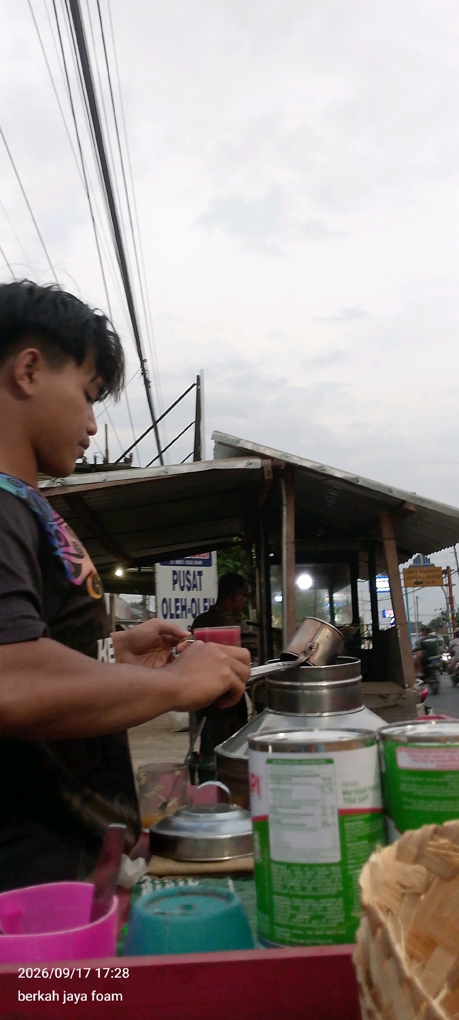 The image depicts a street scene in Kraton, Indonesia, featuring a young man in the foreground preparing a drink. He is wearing a dark t-shirt with a colorful graphic design and holding a glass of pink liquid. In the background, a small food stall with a tin roof is visible, with a sign above it that reads "PUSAT OLEH-OLEH," which translates to "Souvenir Center." Another person is partially visible inside the stall. Various containers, including cans and plastic cups, are arranged on a surface in front of the stall. The environment appears to be outdoors, with power lines and a cloudy sky overhead. A street with moving vehicles is also visible in the background, suggesting an urban setting. The timestamp "2026/09/17 17:28" and the text "berkah jaya foam" are overlaid on the bottom left of the image, potentially indicating the date and location or business name.