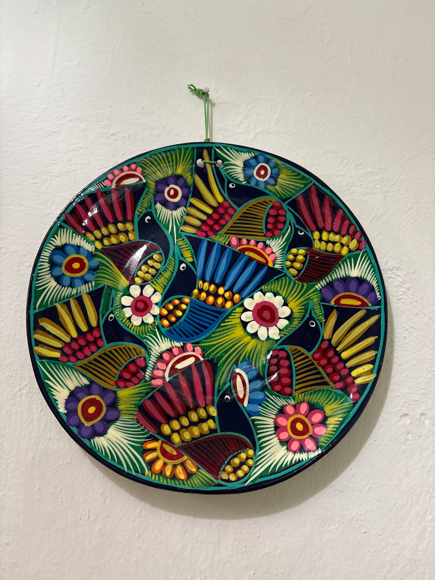 A circular, hand-painted ceramic plate hangs on a textured white wall. The plate features a vibrant, folk-art design with a dark blue background. The design depicts stylized birds with colorful, fan-shaped tails and abstract floral motifs in shades of red, pink, yellow, blue, purple, and green. The plate is suspended by a thin green string attached to a small nail. The image is taken from a medium distance, showing the entire plate and a portion of the wall. No people or other objects are visible. The scene appears to be indoors.
