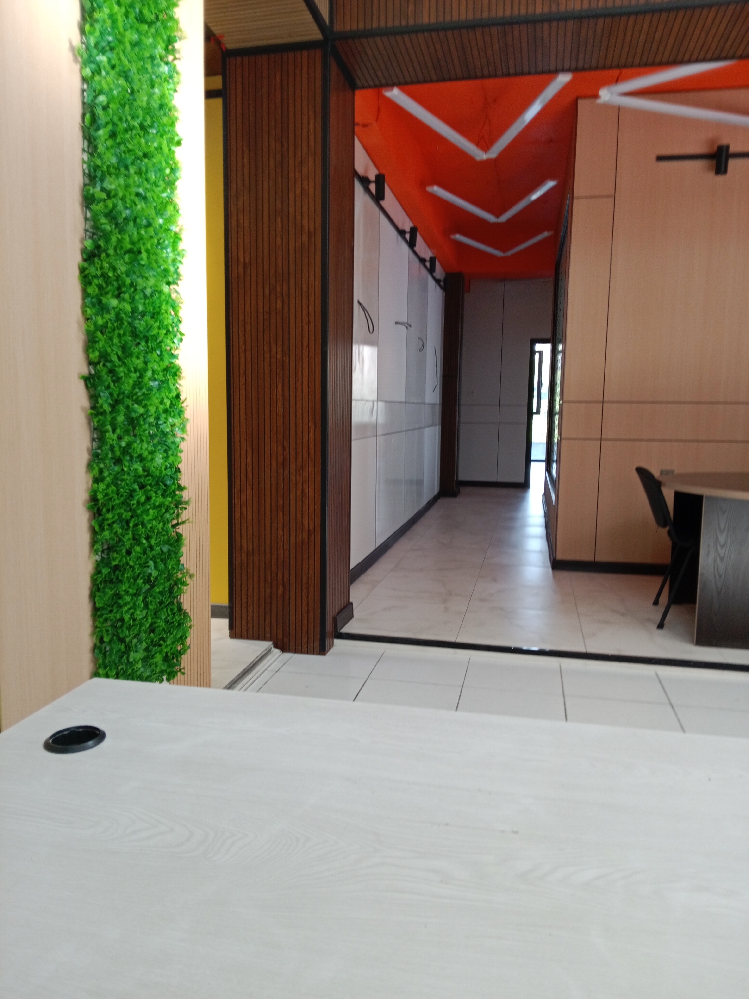 The image depicts the interior of a modern office or co-working space in Tashkent, Uzbekistan. The foreground features a light-colored, wood-grained desk with a black grommet hole. To the left, a vertical section of artificial green foliage adorns a beige wall. Behind the foliage, a bright yellow wall is visible. The middle ground reveals a long corridor with light-colored tiled flooring. On the left side of the corridor are white cabinets with dark trim, and on the right are wood-paneled walls and a dark office chair at a dark desk. The corridor leads to an open doorway. The ceiling of the corridor is an unusual orange color with white angular light fixtures. The overall impression is of a clean, contemporary workspace with some vibrant decorative elements.
