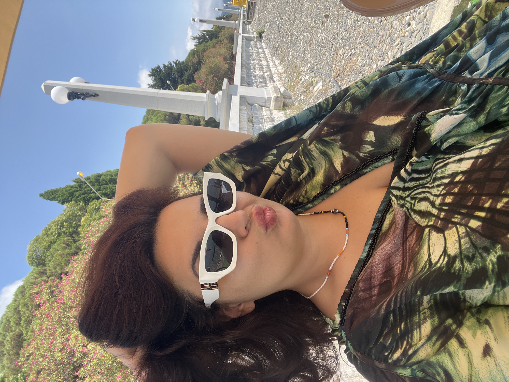 A woman is posing for a selfie outdoors on a sunny day. She has dark, wavy hair and is wearing white sunglasses with a geometric design on the temples and a colorful beaded necklace. Her lips are pursed in a kissing gesture. She is wearing a loose-fitting, flowing garment with a green, brown, and yellow abstract print. The background suggests a coastal location, with a stone wall, a path covered in pebbles, trees, and a clear blue sky. In the distance, there are lampposts and a railing. The environment appears to be a public promenade or park area in Sokhumi, Abkhazia.