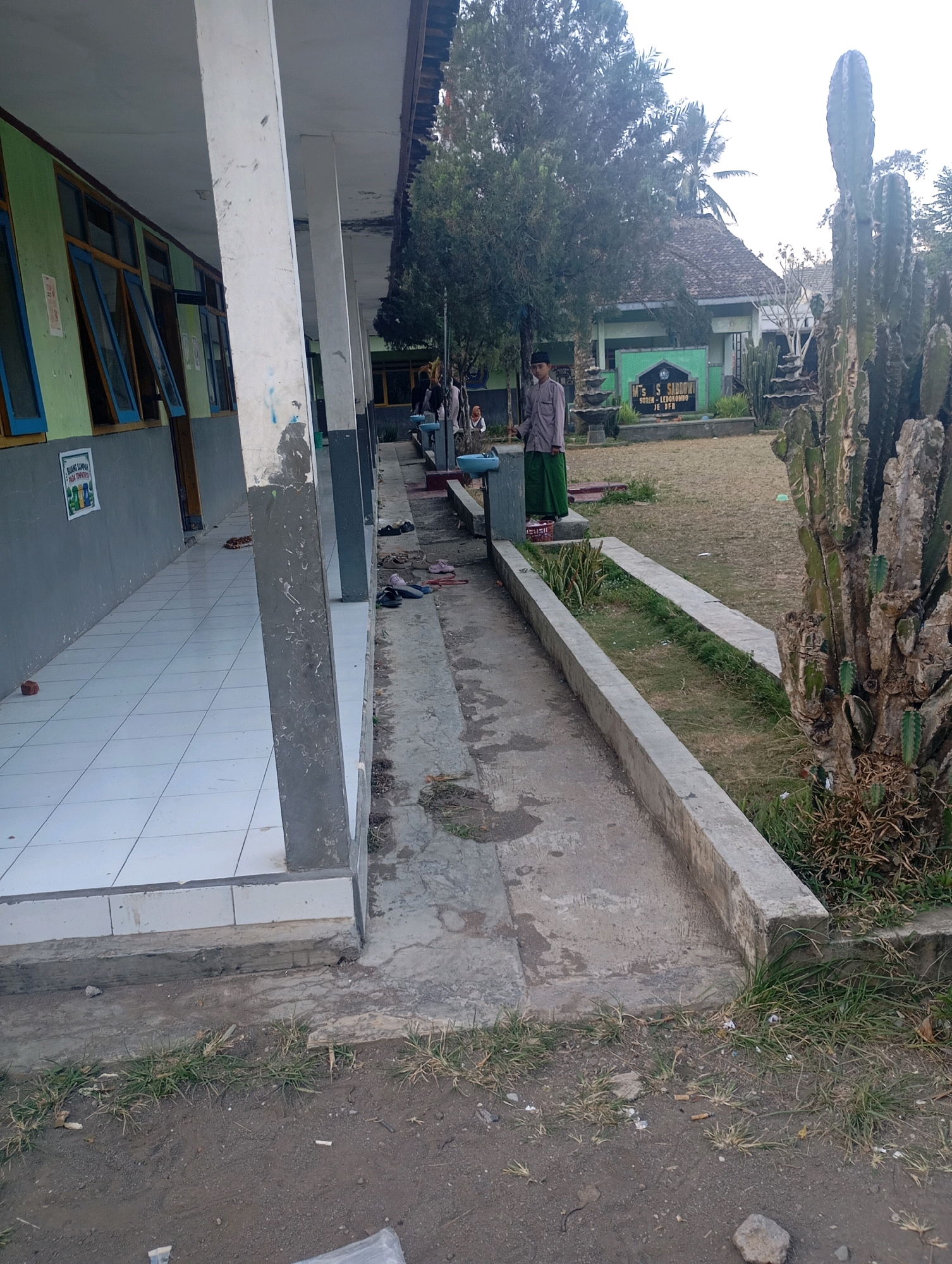 The image depicts an outdoor scene at what appears to be a school in Lembengan, Indonesia. In the foreground, a tiled walkway runs along the side of a building with a covered overhang supported by concrete pillars. The building has green-colored walls and open windows with blue frames. Adjacent to the walkway is a concrete channel with some sparse grass. Further back, a large cactus stands prominently on the right side of the frame. Beyond the cactus, a grassy field stretches towards a building with a tiled roof. A sign is visible in front of this building, displaying text in Indonesian. Several people are present in the scene, including a young man standing in the middle ground wearing a long-sleeved shirt and a green sarong, and another person seated in the background near the building. The ground surface in the foreground is a mixture of dirt and some scattered debris. The overall atmosphere is that of a casual, possibly school-related environment.