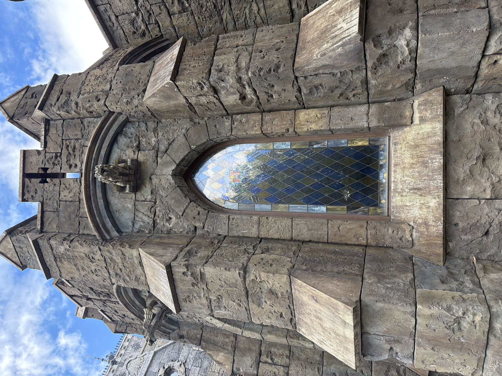 The image depicts a section of a Gothic-style building, characterized by rough-hewn stone blocks and architectural details such as crenellations, a pointed arch window with stained glass, and gargoyle-like sculptures. The stained glass window features a diamond pattern with various shades of blue, yellow, and white, reflecting the sky and surrounding environment. Above the window, a small, grotesque carving resembling a lion or gargoyle is visible. The building's exterior is constructed from large, irregularly shaped stone blocks, giving it an aged and imposing appearance. In the background, a fragment of another building with ornate architectural elements and a spire can be seen against a blue sky with scattered clouds. The scene appears to be an outdoor shot focusing on the facade of the building. The location in Khryashchevka, Russia, likely refers to the Khryashchevka Palace of Culture, which is known for its unique architectural style.