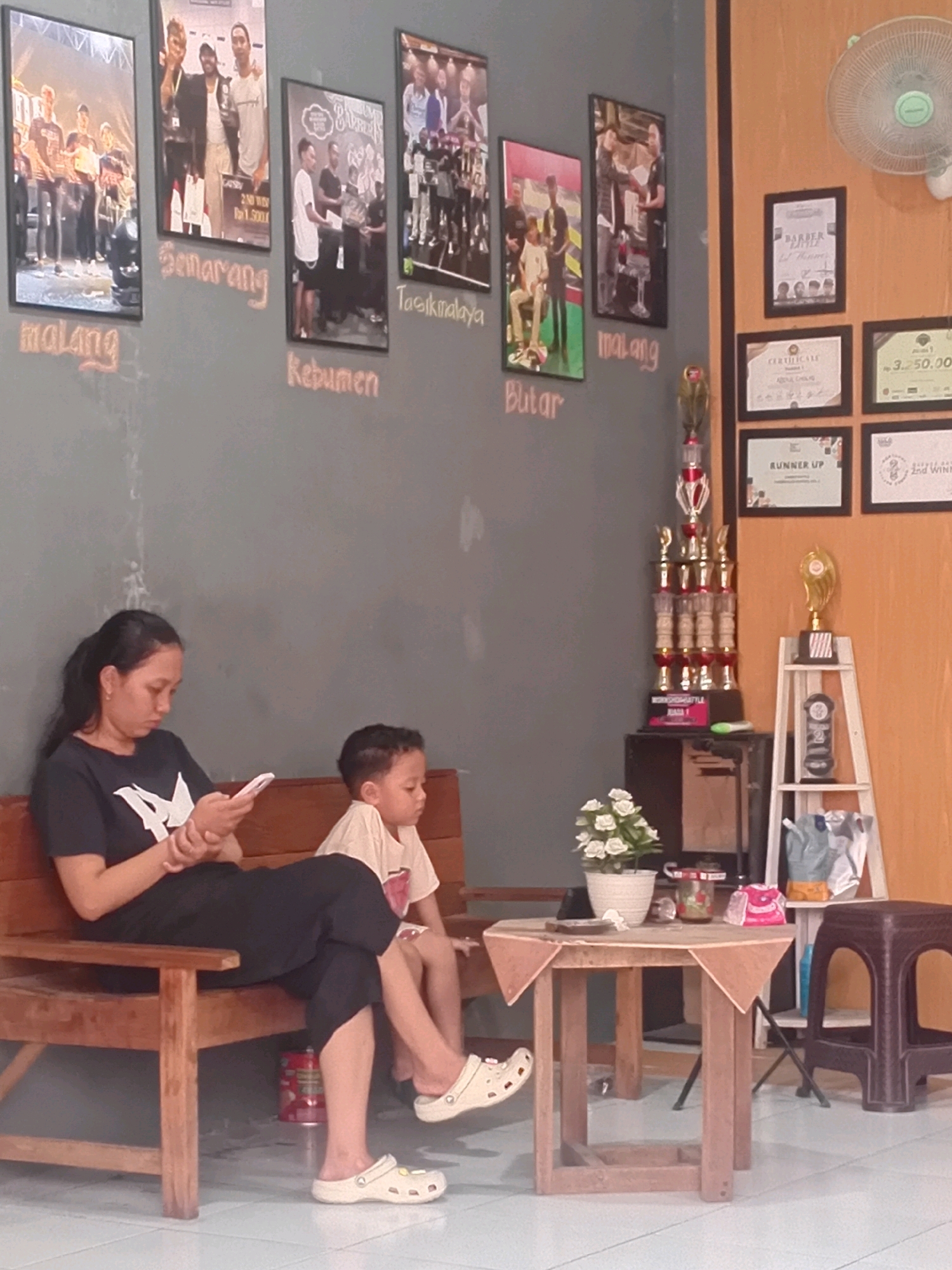 This is a medium shot of a woman and a young boy sitting on a wooden bench in what appears to be a waiting area. The woman is on the left, wearing a black t-shirt and black pants, and is looking down at her white smartphone, holding it with both hands. The boy is to her right, wearing a light-colored t-shirt and dark shorts, and is looking forward. Both are wearing casual shoes.
The wall behind them is a dark grey and is decorated with several framed photographs and certificates. The photographs depict groups of people, some holding trophies, and appear to be related to competitions or events. Text in stylized lettering is present below some of the photos, including "Malang", "Semarang", "Kebumen", "Tasikmalaya", and "Blitar", which are cities in Indonesia. To the right of the photographs, there is an orange wall section with a white oscillating fan mounted high up. Below the fan, a series of framed certificates and a single gold trophy on a white display shelf are visible. A tall, multi-tiered trophy is also present on a dark surface to the left of the display shelf.
In the foreground, to the right of the bench, is a small, square wooden table covered with a tan-colored tablecloth draped over its corners. On the table, there is a white pot with white flowers, a clear jar with a red lid, and a pink and white object. A dark brown plastic stool with arched openings is positioned to the right of the table. The floor is tiled in a light blue and white pattern. The overall impression is of a waiting area, possibly at a community center or competition venue, in the city of Besito, Indonesia.