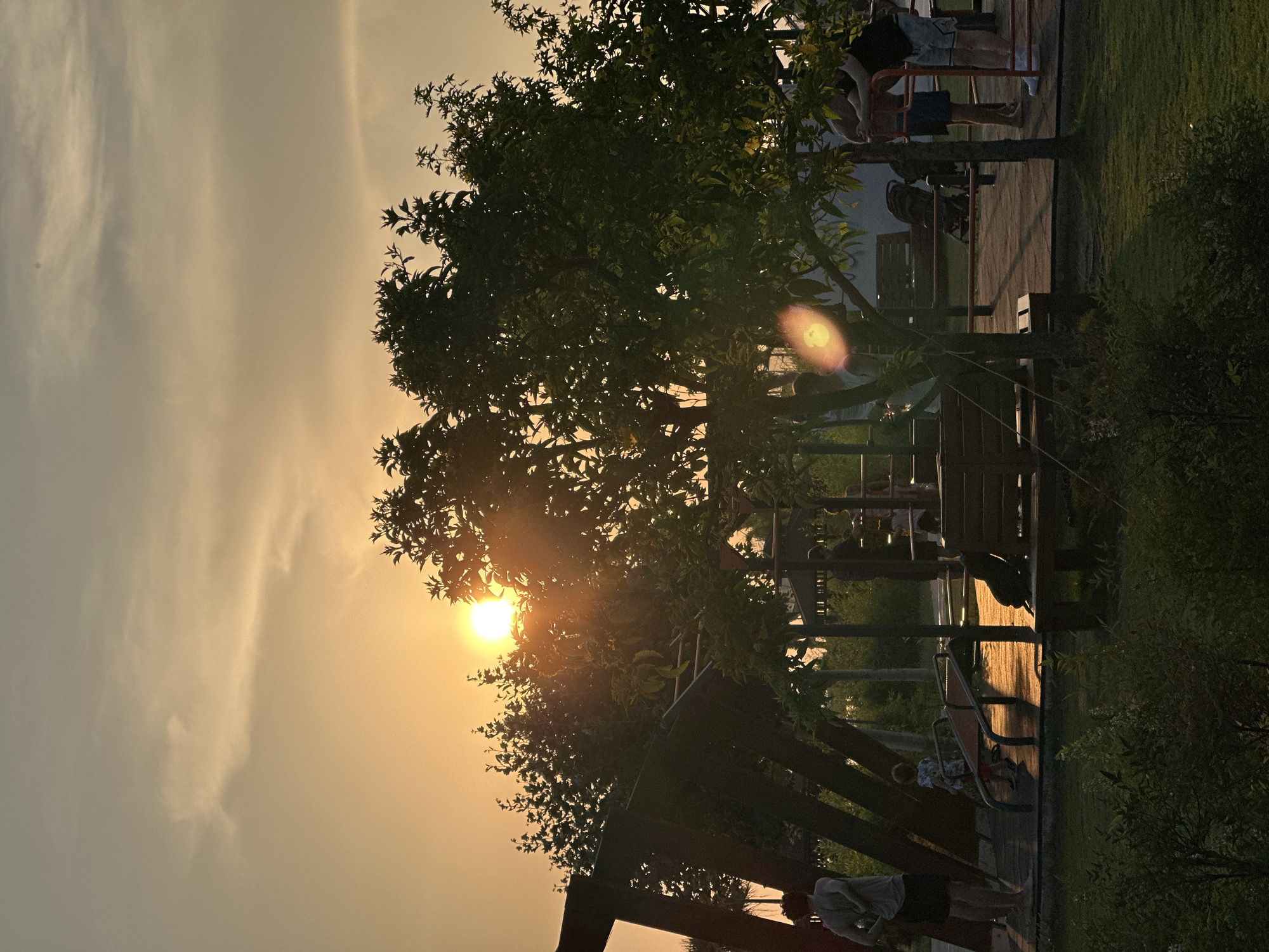 The image depicts a park scene at sunset in Fethiye, Turkey. The sun is setting on the left side of the frame, casting a warm orange glow across the sky and illuminating a portion of the playground equipment. Several trees with dark green foliage are prominent in the foreground and middle ground, partially obscuring the sun. Silhouetted figures are visible throughout the park. On the left, a person stands with their back to the camera, wearing a gray t-shirt and dark shorts. Nearby, a child in a patterned shirt and red shorts is standing on a playground structure. In the middle ground, a wooden bench faces away from the viewer, and further back, two individuals stand near some red-railed equipment. A stroller is also visible in the middle ground. The ground of the playground appears to be a dark, reddish-brown surface, with patches of green grass in the foreground and background. The overall atmosphere is serene, with the setting sun creating a peaceful ambiance.