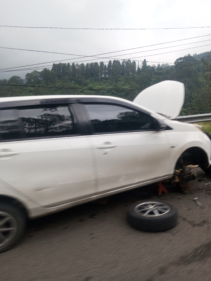 The image displays a white passenger vehicle, possibly a sedan or compact MPV, situated on an asphalt road in Jabalpur, India. The vehicle is elevated on a red jack stand beneath its front passenger side, where the wheel has been removed, exposing the brake disc and caliper assembly. A single wheel with a silver-colored rim lies flat on the road surface directly in front of the elevated section. The vehicle's hood is open. Scattered metallic objects, possibly tools, are visible near the detached wheel. The immediate background shows a metal guardrail, beyond which a dense, verdant hillside covered with numerous trees rises. The sky is overcast and grey. Multiple dark utility cables traverse horizontally across the upper portion of the frame. No individuals are visible in the scene.