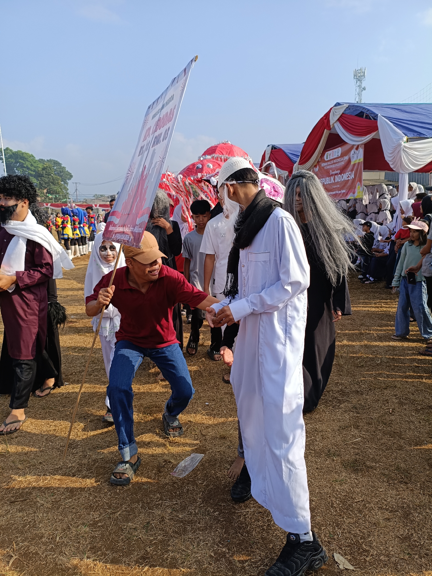 The image shows an outdoor event, likely a parade or cultural festival, in Cibodas, Indonesia. A man in a red shirt and blue jeans is holding a long stick with a white banner attached, interacting with a person dressed in white with a black scarf. Other individuals are present, some in costumes, including one with long grey hair and another with a black beard and white scarf. In the background, there are tents with red and white drapes and a banner that reads "REPUBLIK INDONESIA". A group of people, some in uniform, are visible in the distance. The ground is covered with dry grass and dirt. The sky is clear and blue.