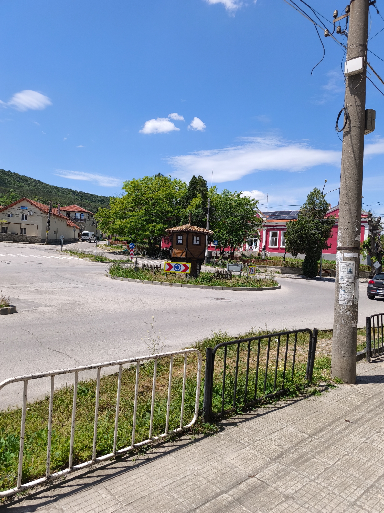 The image captures a roundabout in an unidentified town or city. A small, wooden structure resembling a miniature house sits in the center of the roundabout, surrounded by greenery and road signs indicating the direction of travel. The surrounding buildings consist of residential houses and a prominent red building, possibly a public structure, fitted with solar panels on its roof. In the foreground, a white and black metal railing runs along the edge of the sidewalk, casting shadows on the tiled pavement. A concrete utility pole stands adjacent to the railing, adorned with wires and various attachments. The sky is clear blue with a few scattered white clouds, suggesting a sunny day.