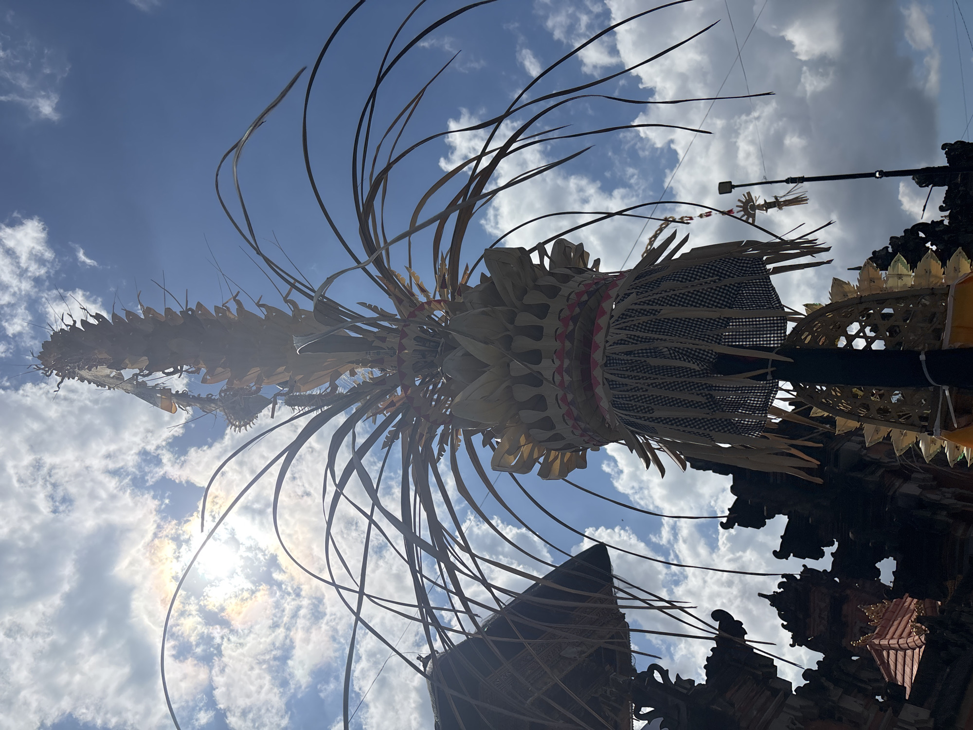 In Mengwi, Indonesia, a tall, ornate structure, likely a ceremonial offering or decoration, stands against a bright blue sky dotted with white clouds. The structure is adorned with dried leaves and woven materials, giving it a layered, textured appearance. Its upper portion tapers to a point, while the lower section features decorative fringe and intricate weaving. To the left, a portion of a traditional Balinese building with a thatched roof is visible, suggesting a temple or cultural site. The sun is partially obscured by clouds, casting a slight rainbow effect in the upper left corner of the image. The scene is captured from a low angle, looking up at the sky and the elaborate structure.