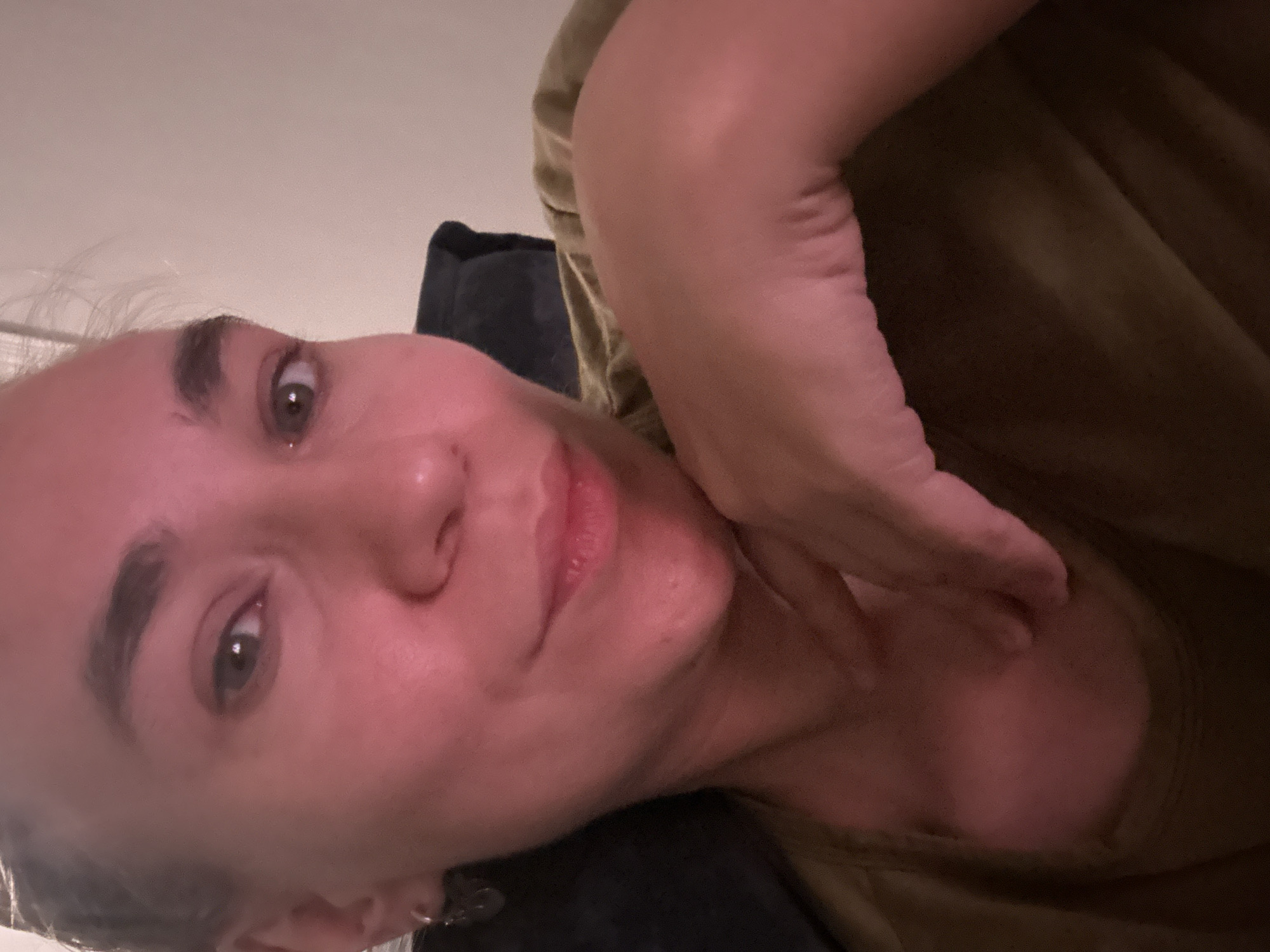 A close-up, angled headshot depicts a person with light skin and graying hair. The person's left hand is near their neck. The person is wearing a dark-colored cushion on their head and a brown top. The image does not contain information to infer the location.