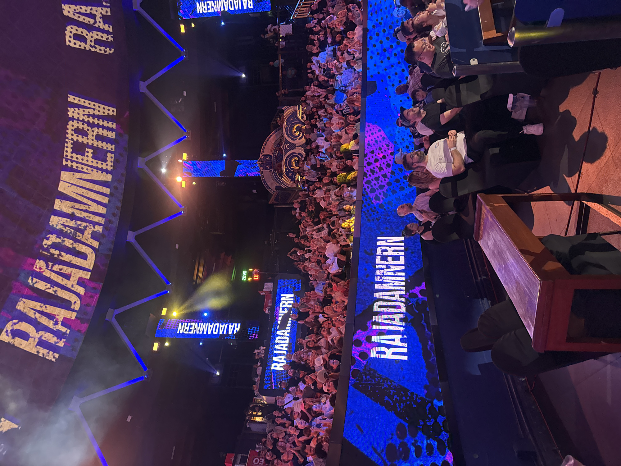 The image depicts a densely packed indoor arena in Bangkok, Thailand, filled with spectators. Bright displays prominently show the word "RAJADAMNERN." Stage lighting and structural beams are visible above the crowd. Some people are seated, while others are standing. The scene is dimly lit except for the brightly lit screens and stage area.