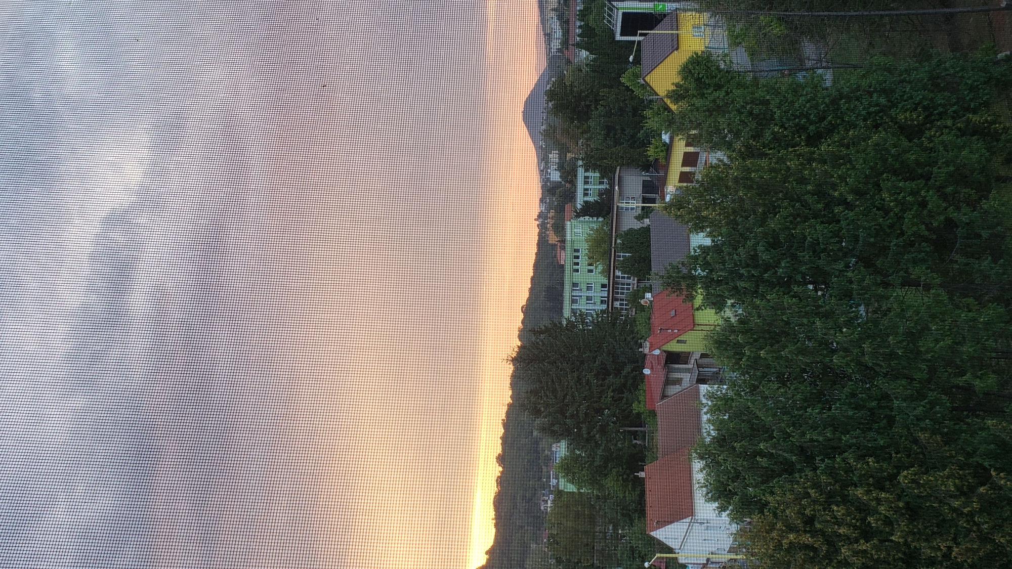 This image depicts a view through a window screen of a suburban area in Prešov, Slovakia, during what appears to be sunset. The sky is a gradient of soft orange and grey clouds. In the mid-ground, there are several residential buildings with red and grey tiled roofs, interspersed with lush green trees. Further in the background, a larger, light green building complex is visible, likely an apartment building or institutional facility. Beyond that, a hill covered in trees dominates the horizon, with a distant peak visible to the right. The overall impression is one of a tranquil, residential neighborhood.