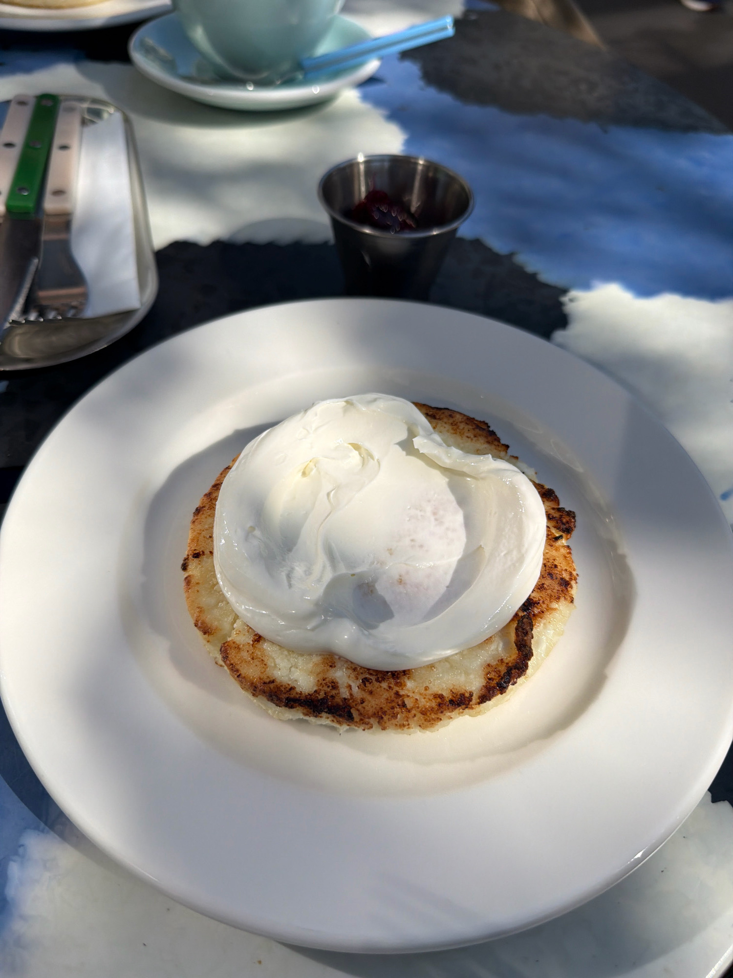 A close-up shot shows a single serving of food on a white plate. The dish consists of a round, toasted bread or pancake topped with a perfectly poached egg. The egg white is fully cooked and encases a visible yolk. The plate is positioned on a table with a patterned blue and white surface, casting a shadow from the food. To the left, a metal utensil holder with a knife and fork with green and beige handles is visible. Further back, a small metal ramekin contains a dark red preserve, likely jam. A blurred light blue cup and saucer with blue straws are also in the background, suggesting a cafe or restaurant setting. The lighting is bright and appears to be natural, casting distinct shadows. The scene does not contain any identifiable individuals or provide specific clues about the location within Moscow, Russia.